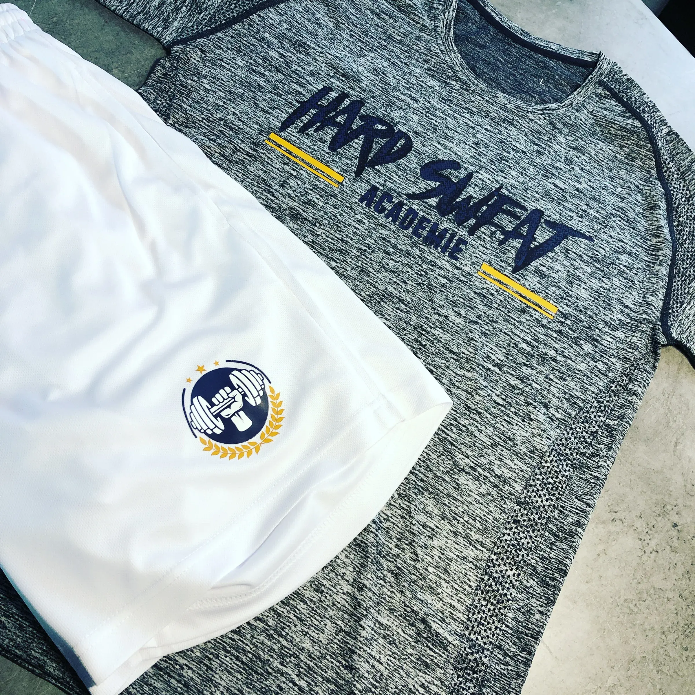 Maillot gris chiné et short blanc marqués d'un lettrage bleu marine et d'un logo doré, posés à plat