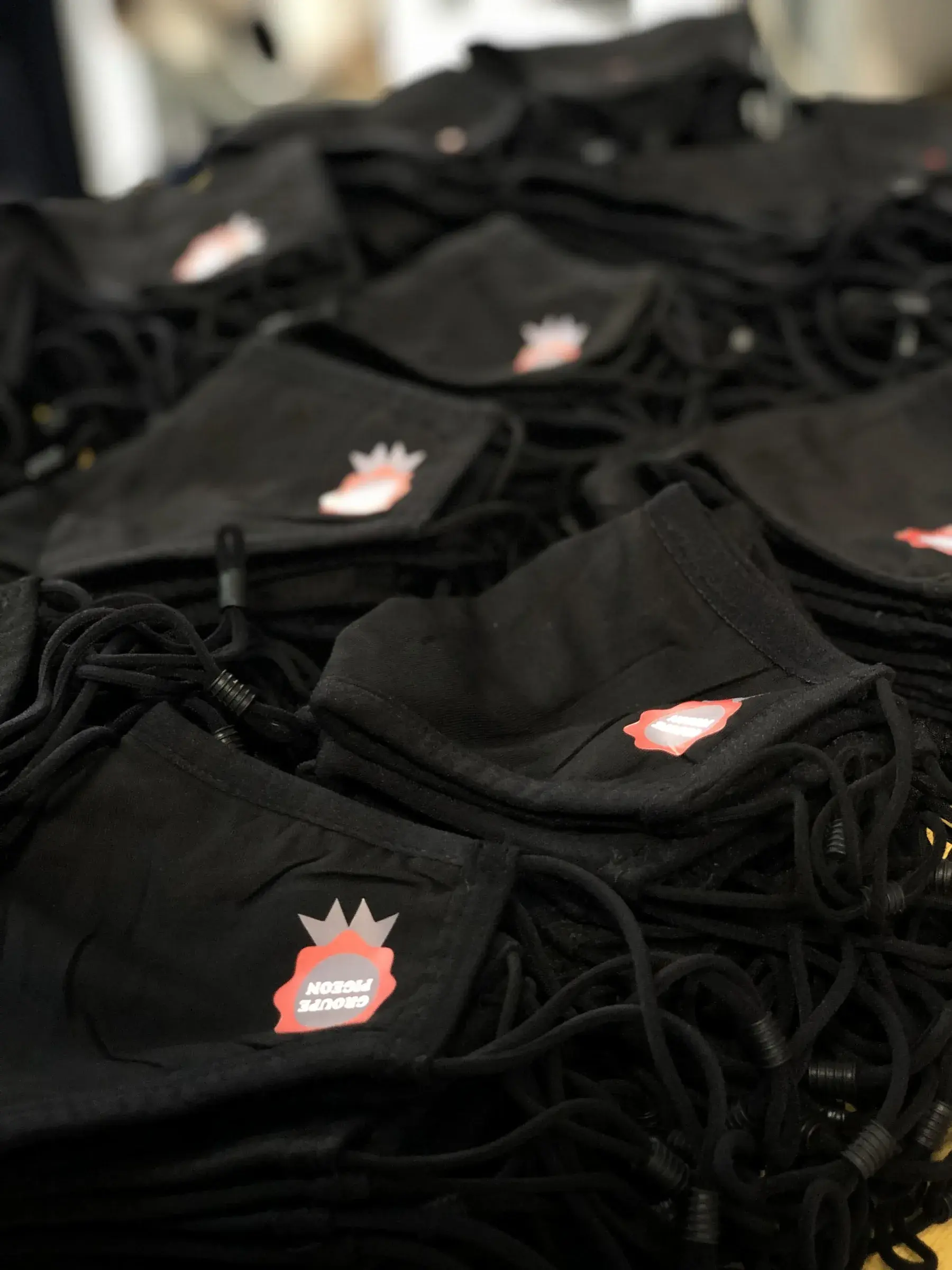 Masques en tissu noir marqués d'un petit logo rouge et blanc, empilés en grande série avec leurs cordons