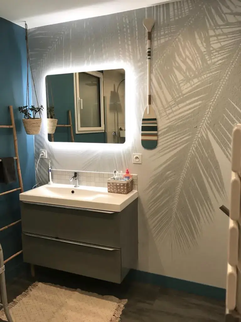 Salle de bain avec un mur habillé d'un papier peint à feuilles de palmier gris et blanc, miroir rétroéclairé et meuble vasque gris