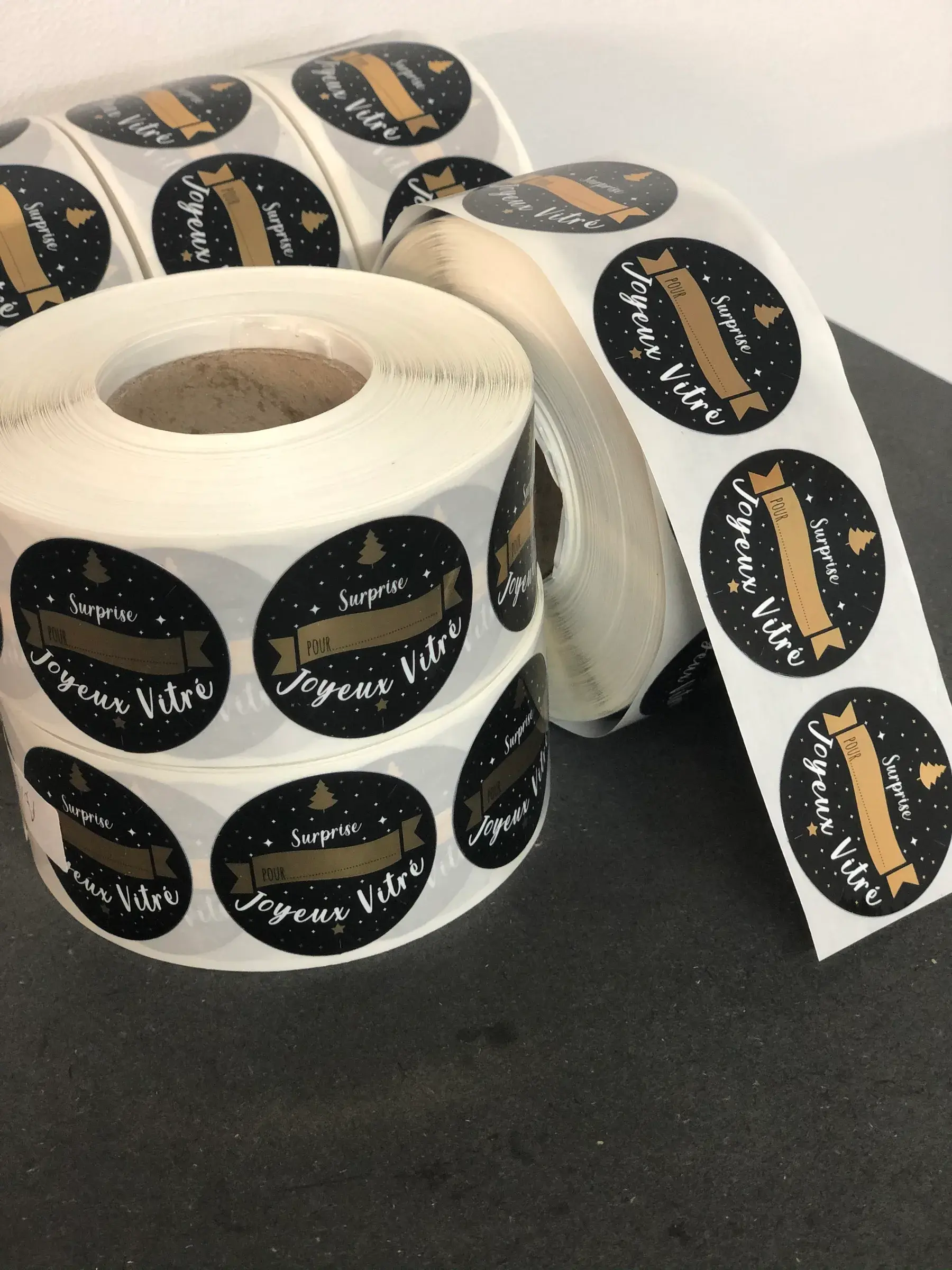 Rouleaux d'étiquettes rondes noires à ruban doré et lettrage blanc sur le thème de Noël, posés sur une table