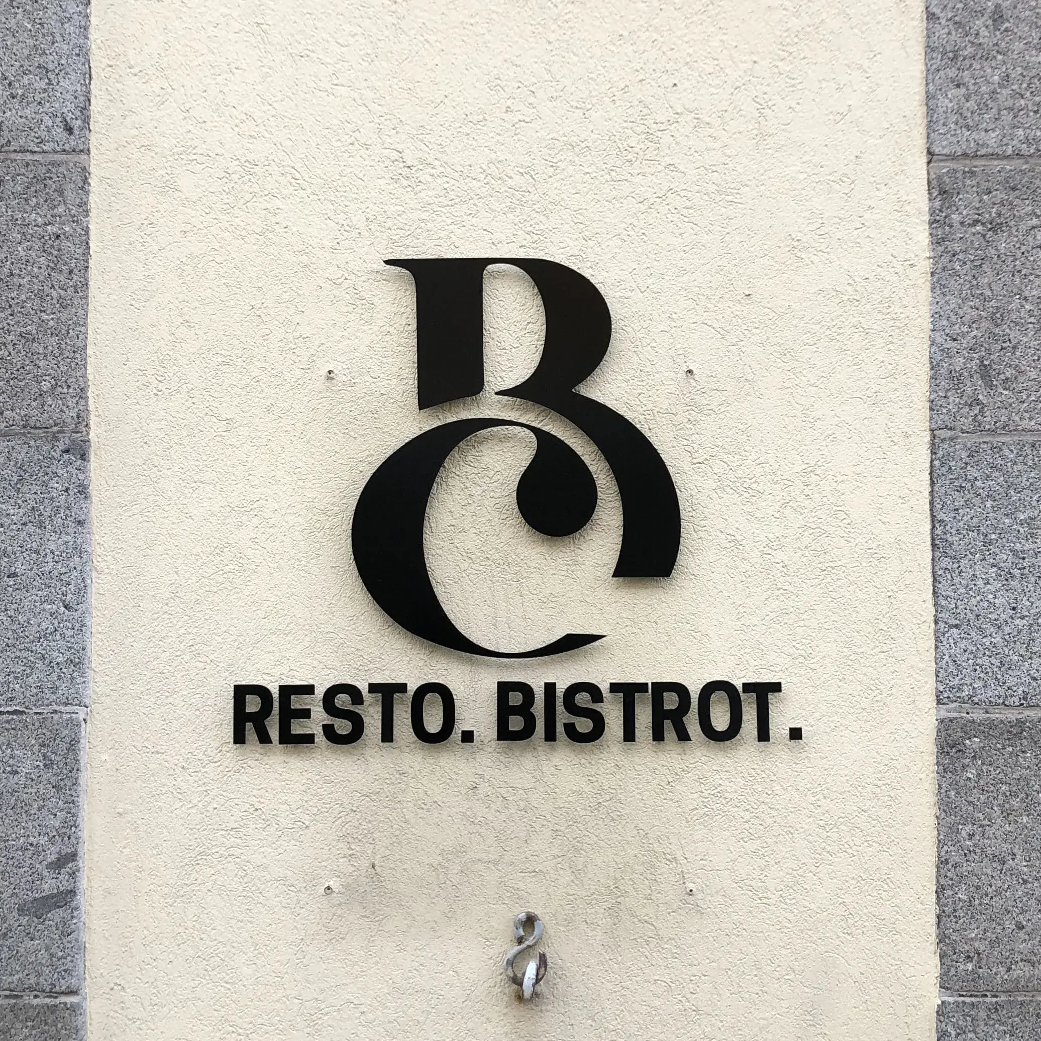 Monogramme et lettres noires découpées en relief sur un mur crépi clair, entre deux piliers de granit