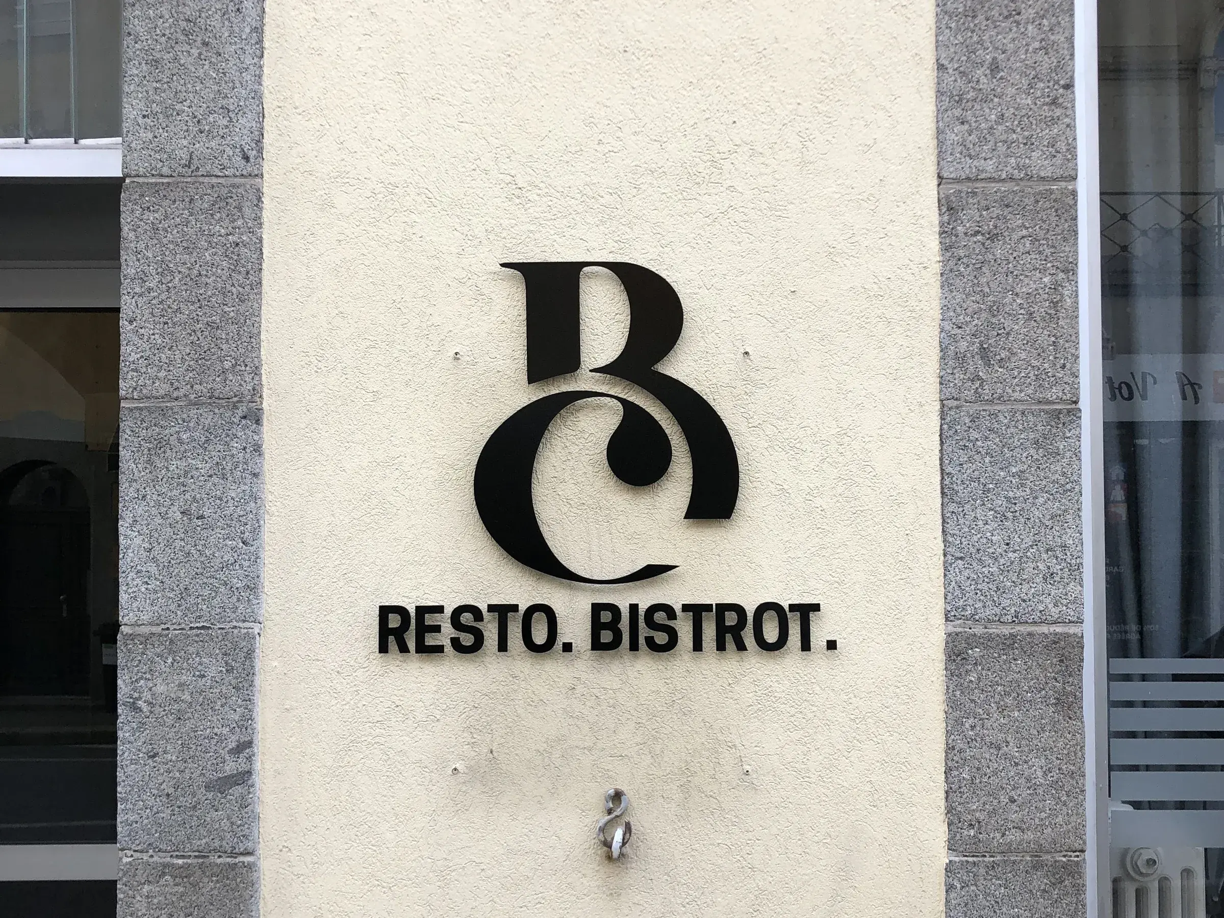 Monogramme et lettres noires découpées, posés en relief sur le mur crépi d'un restaurant entre deux piliers de granit