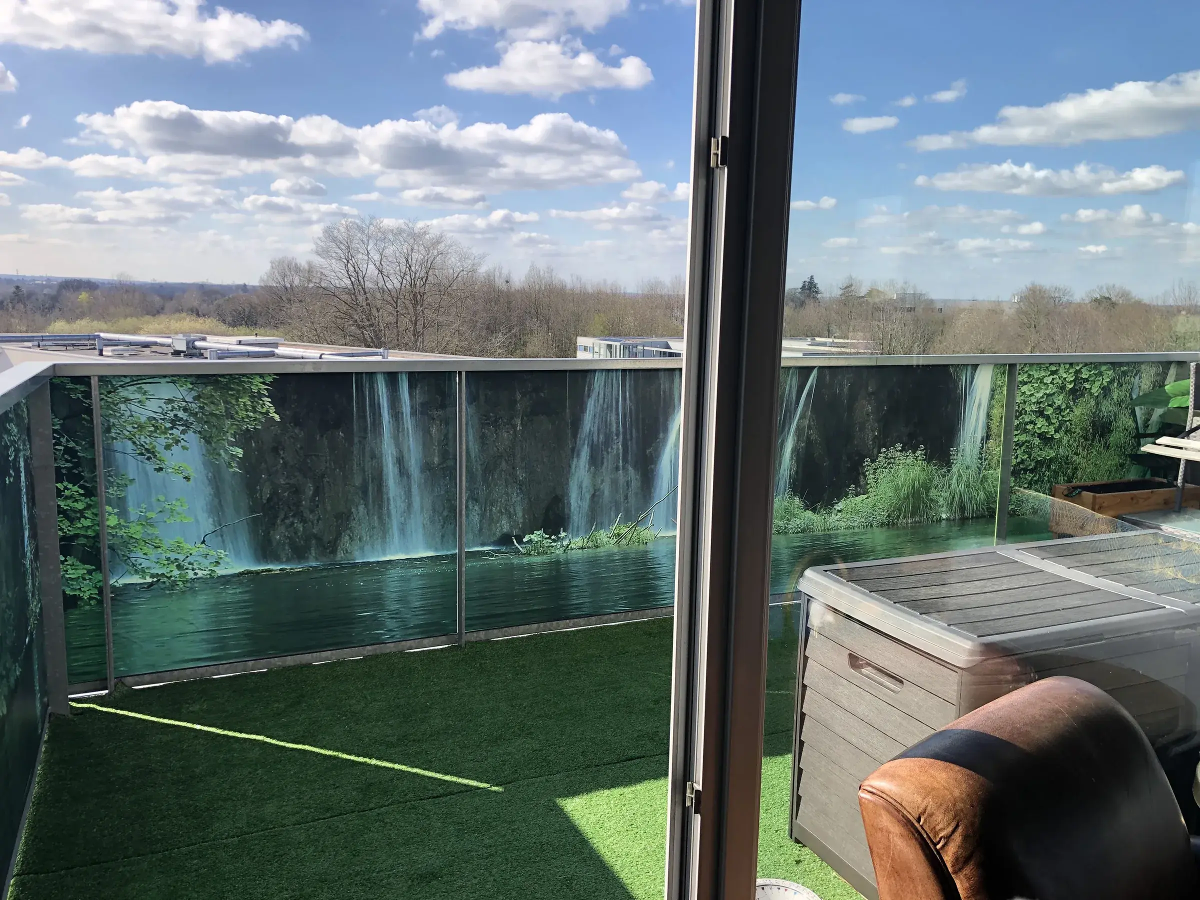 Balcon dont les garde-corps vitrés sont habillés d'un visuel imprimé de cascades et de végétation, gazon synthétique au sol