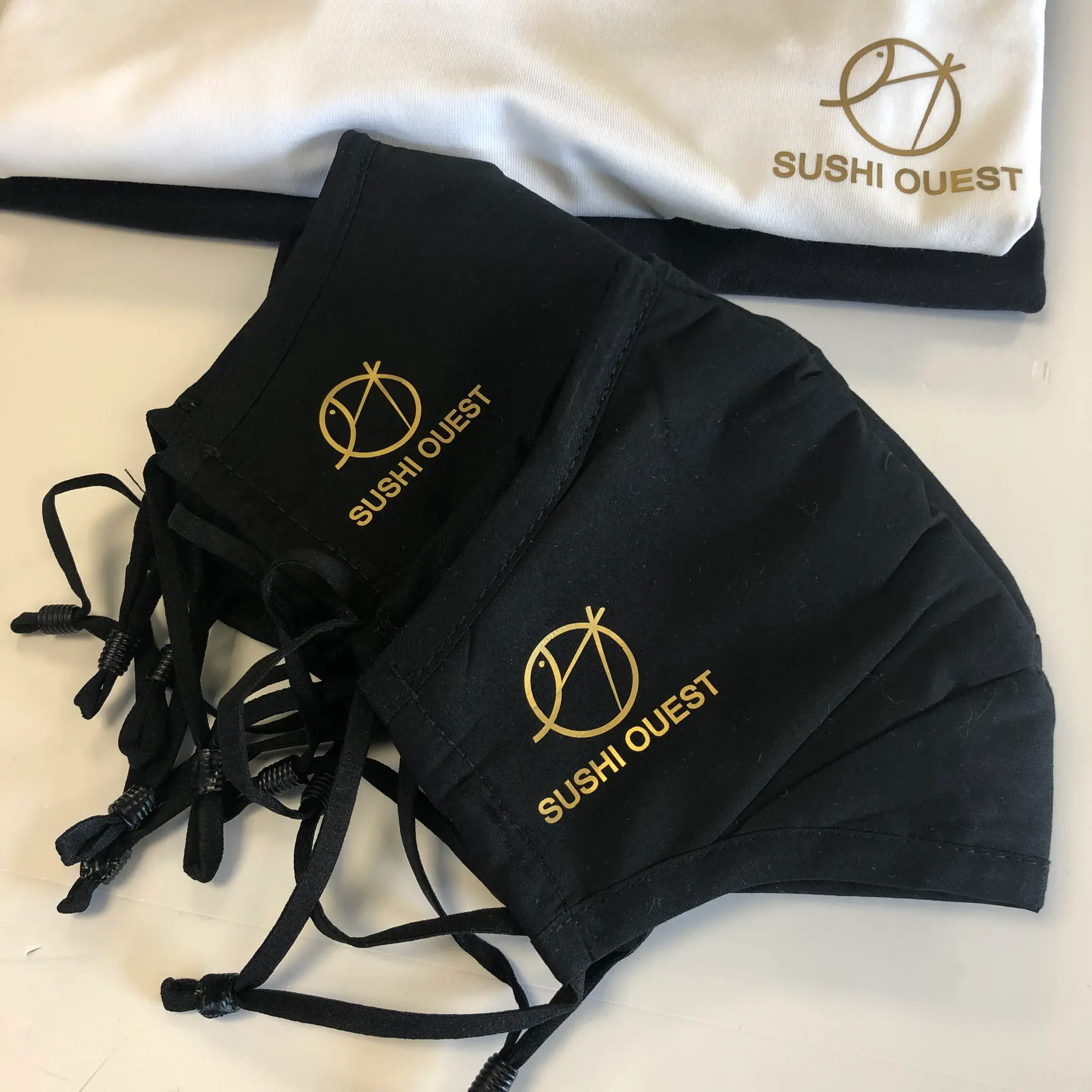 Masques en tissu noir imprimés d'un logo doré, posés sur un tee-shirt blanc marqué du même logo