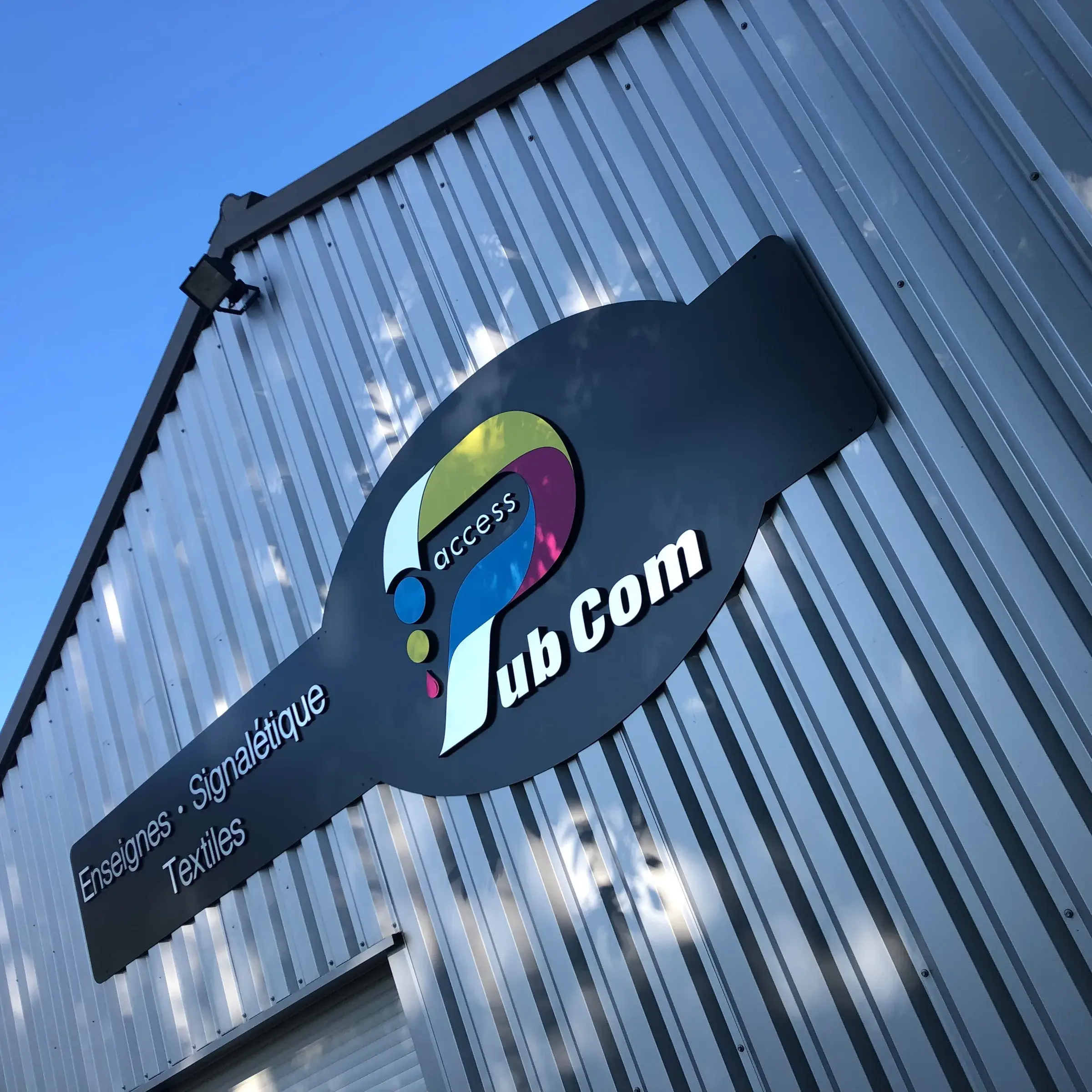 Enseigne en relief au logo d'Access Pub Com sur le bardage métallique de l'atelier, en contre-plongée sous un ciel bleu