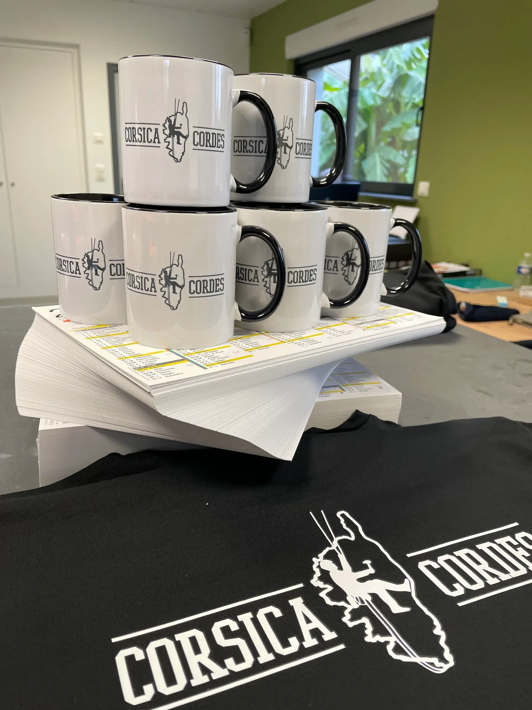 Mugs blancs à anse noire marqués d'un logo noir, empilés sur des imprimés, avec un tee-shirt noir sérigraphié au même logo au premier plan