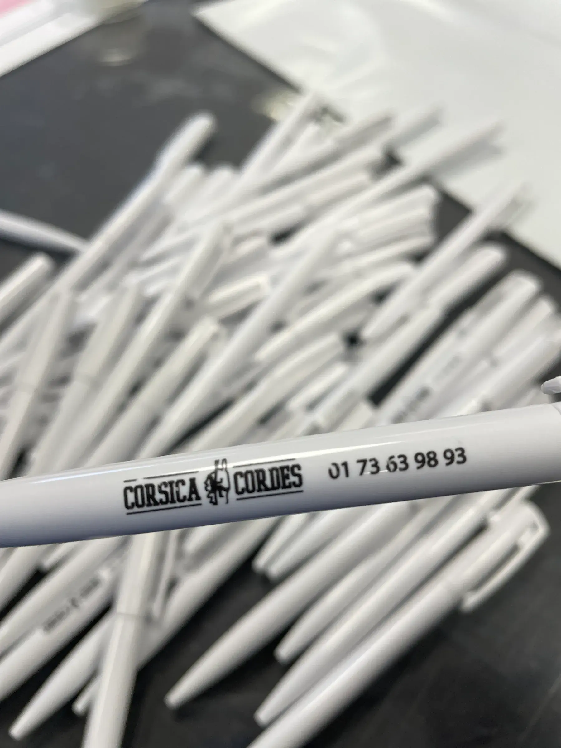Stylos blancs marqués d'un logo et d'un numéro de téléphone en noir, étalés en vrac sur une table sombre