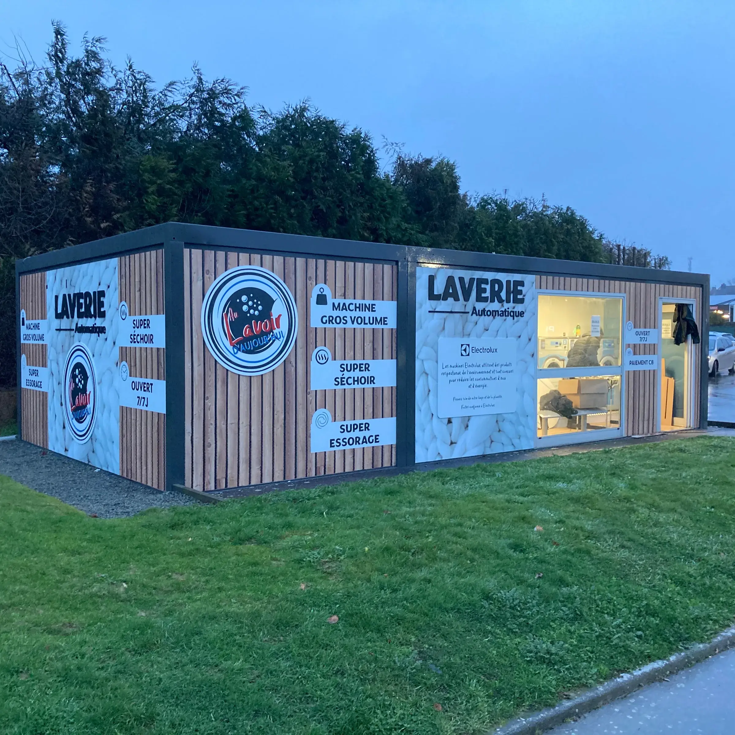 Laverie automatique en conteneur habillée de bois et de visuels de linge, vitrine éclairée à la tombée du jour
