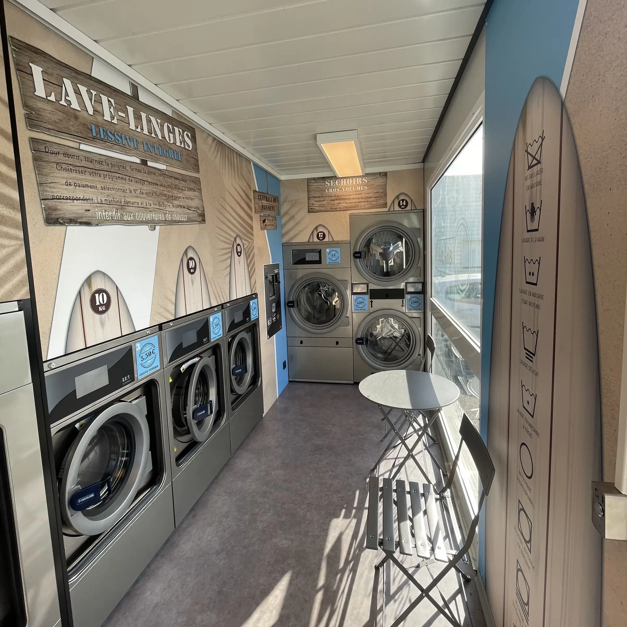 Intérieur d'une laverie habillé de décors imprimés sur le thème du surf, machines alignées et table de bistrot près de la baie vitrée