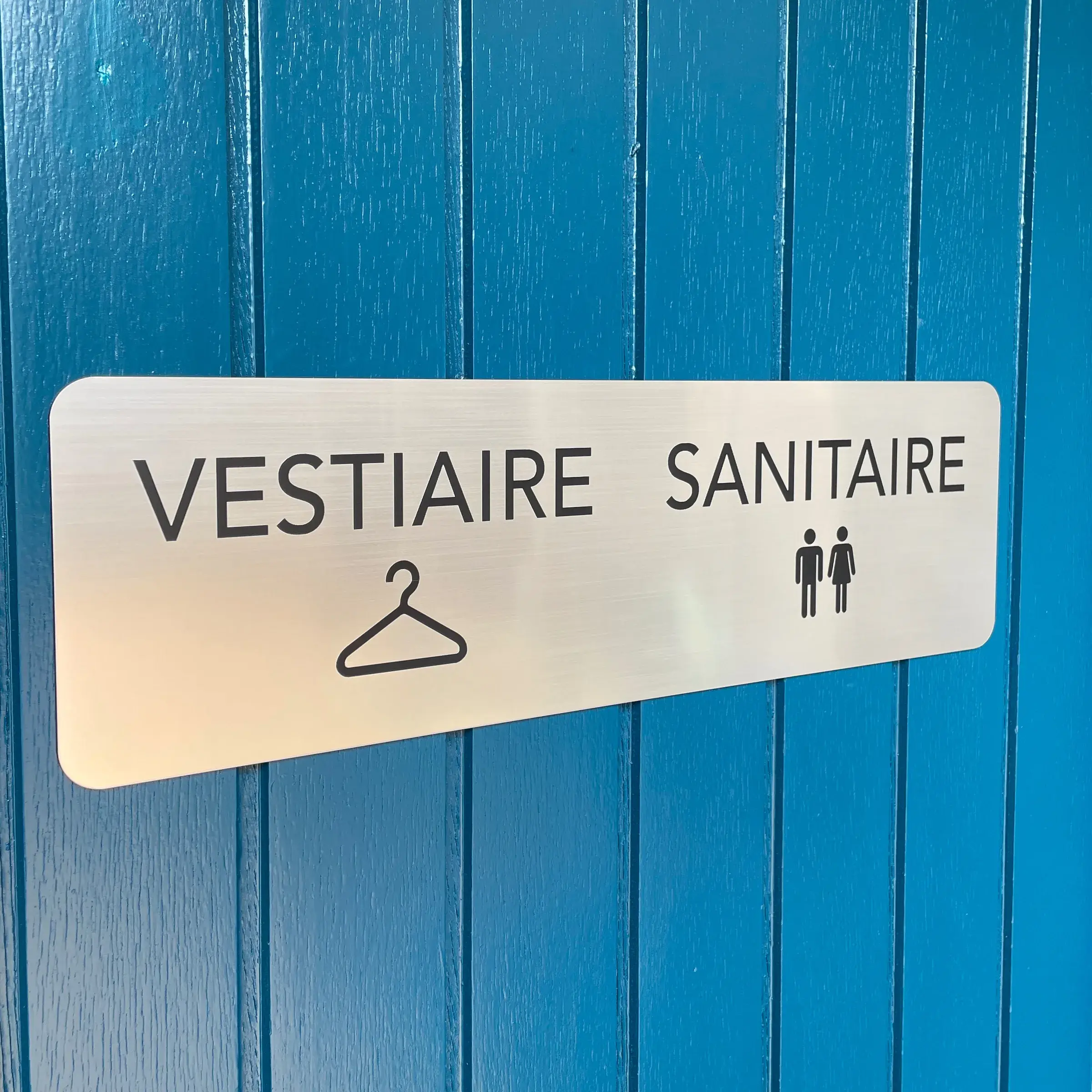 Plaque en aluminium brossé avec pictogrammes de vestiaire et de sanitaires, fixée sur un mur en lambris bleu