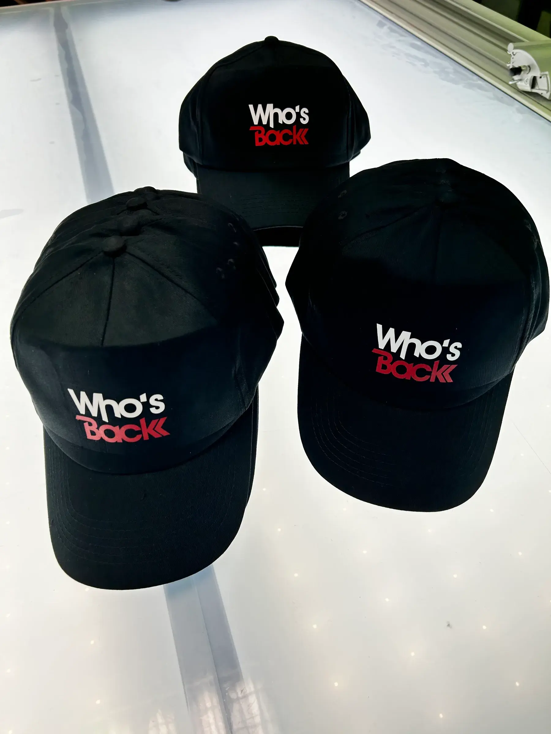 Trois casquettes noires imprimées d'un logo blanc et rouge, posées sur une table lumineuse