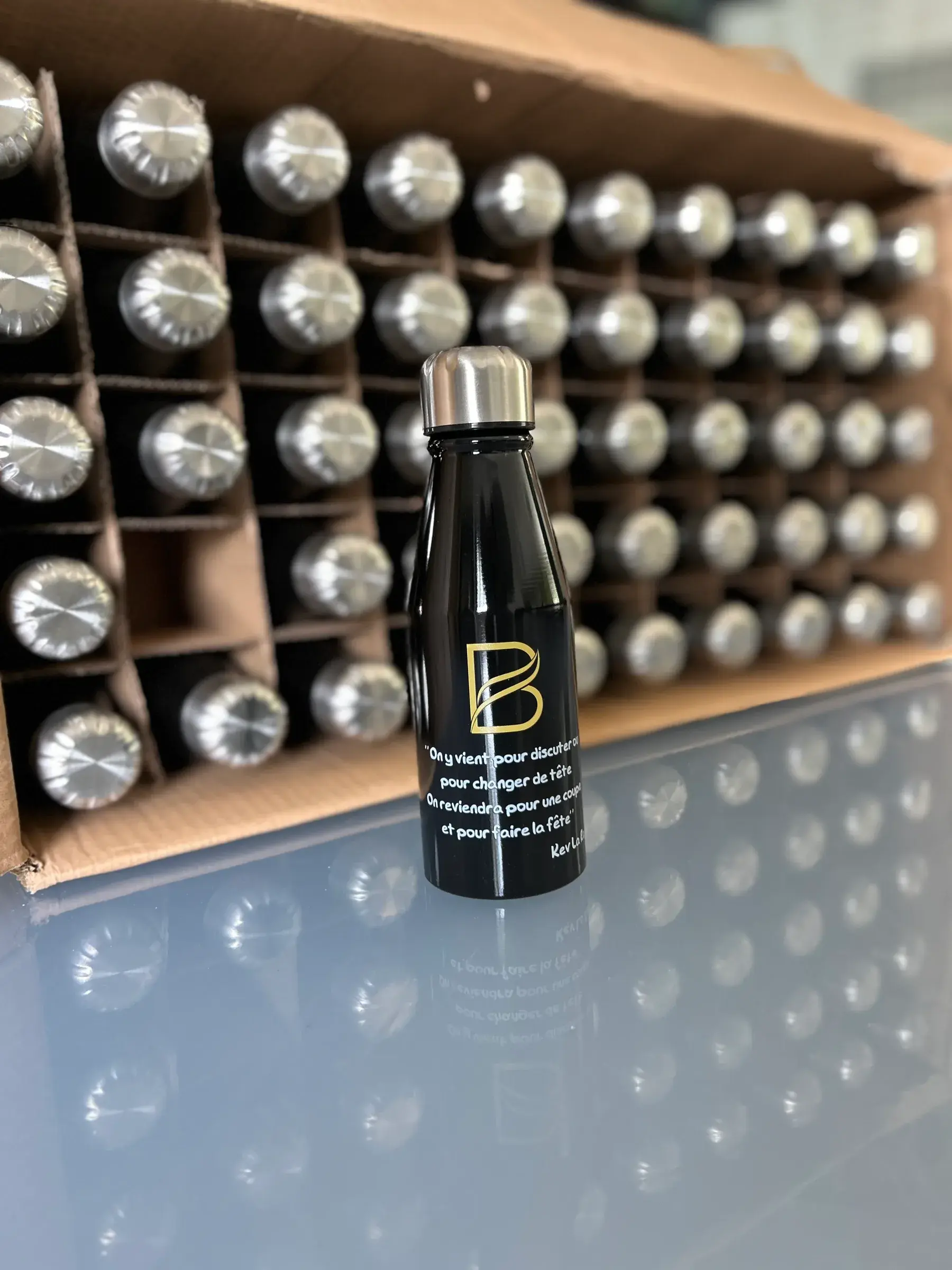 Bouteille isotherme noire marquée d'un monogramme et d'une citation dorés, devant un carton de bouteilles identiques