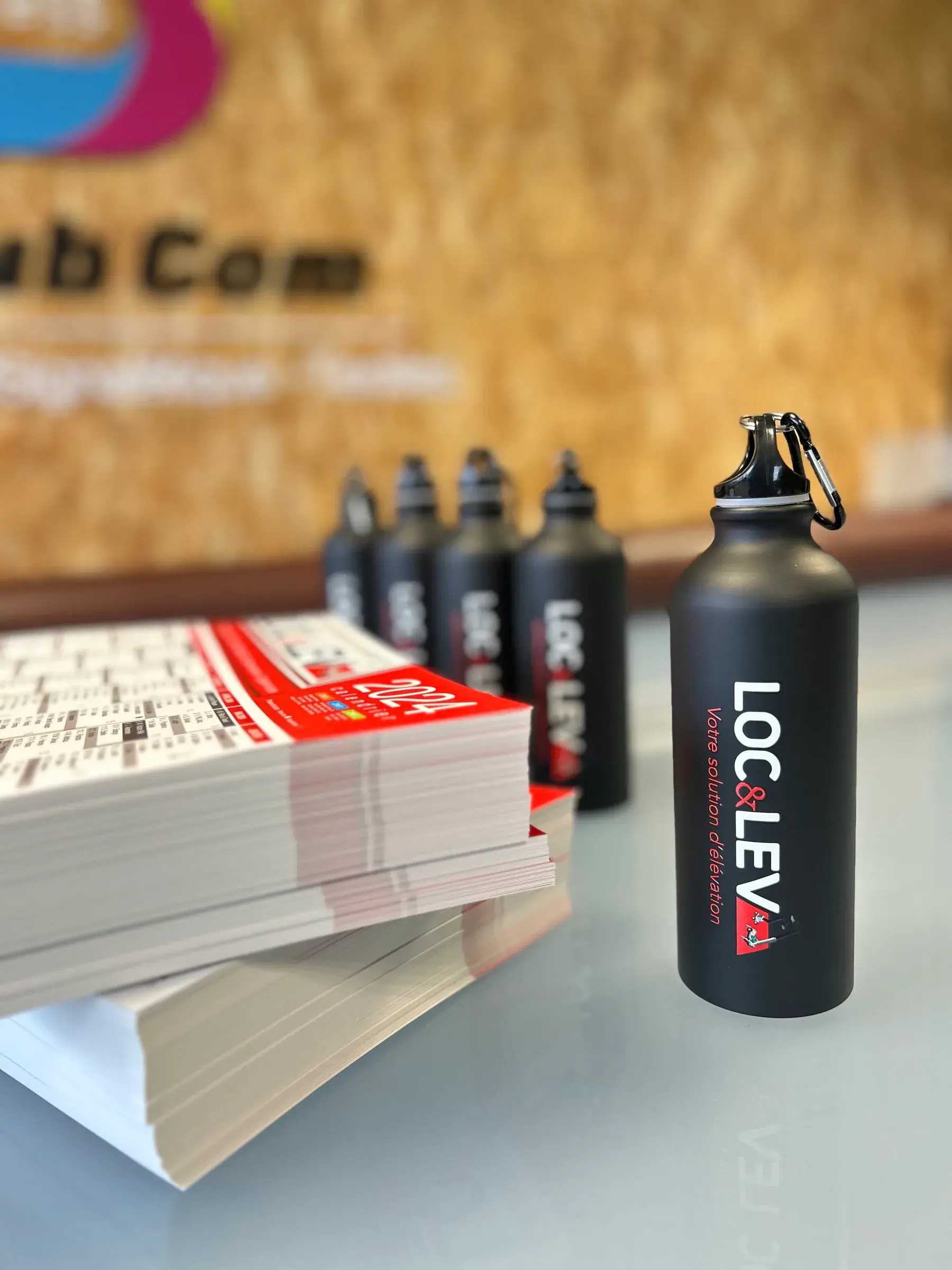 Gourdes en aluminium noir marquées d'un logo blanc et rouge, à côté d'une pile de calendriers, devant le panneau au logo d'Access Pub Com