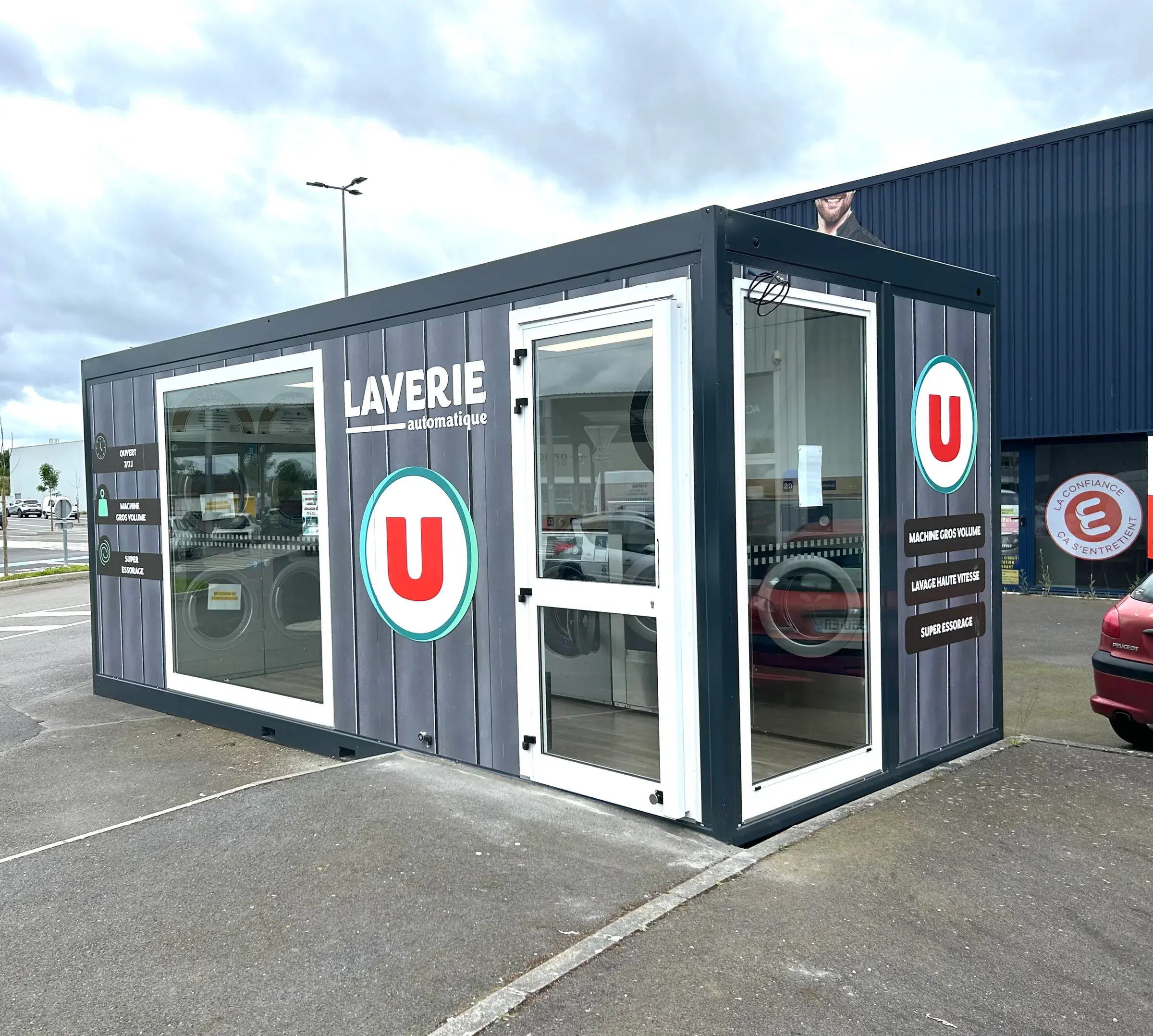 Laverie automatique en conteneur habillée d'un décor gris avec logos ronds sur les flancs, sur le parking d'un magasin