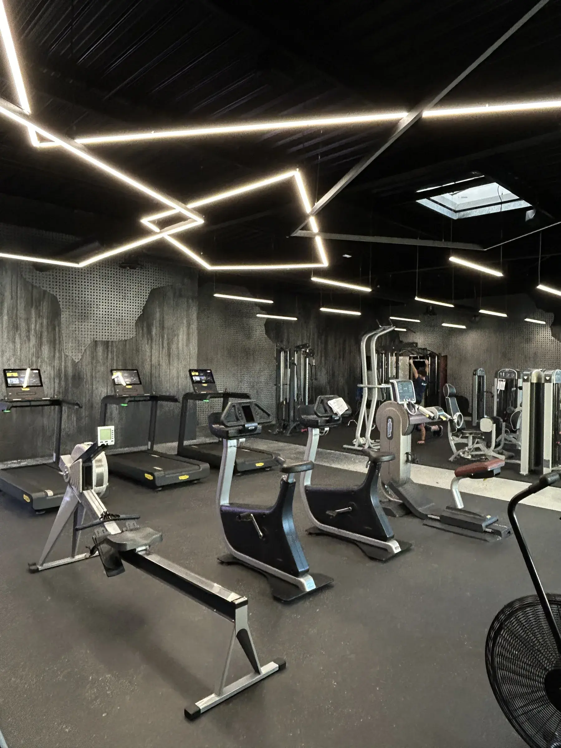 Salle de sport aux murs habillés d'un décor imprimé effet béton et métal perforé, lignes lumineuses au plafond au-dessus des appareils de cardio