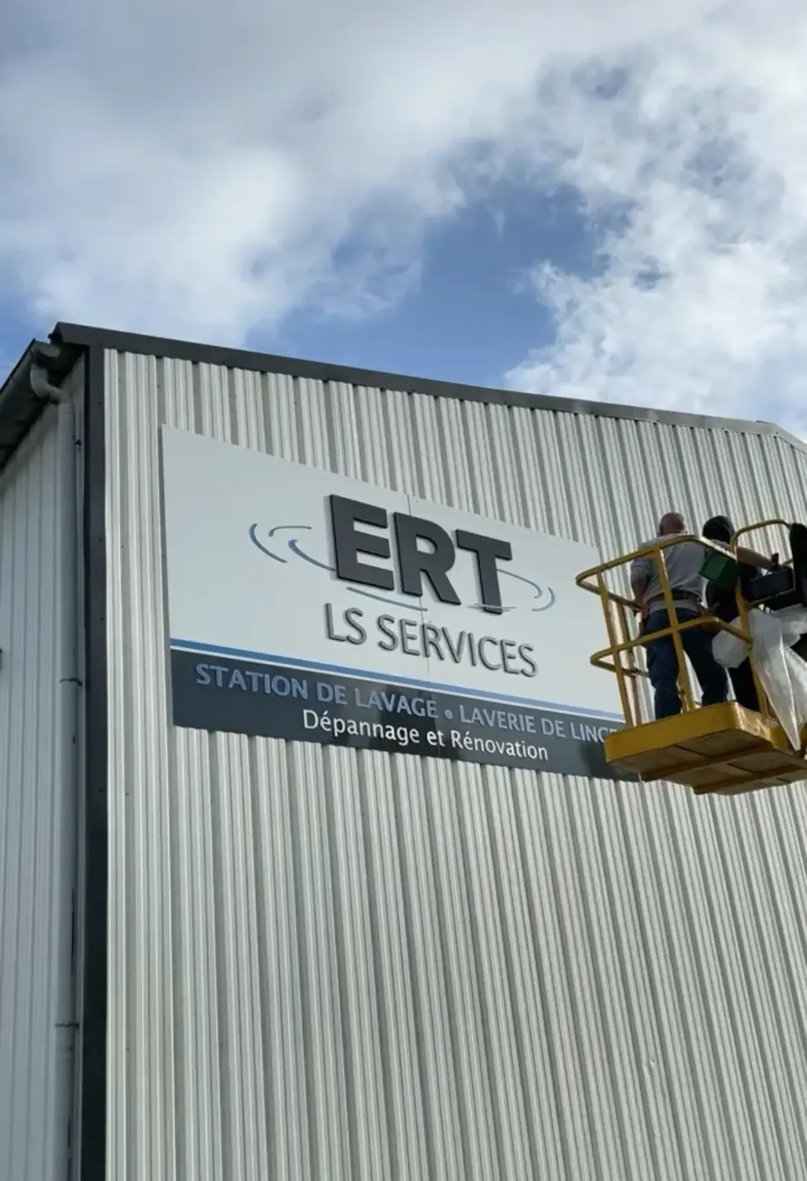 Deux poseurs en nacelle fixant un grand panneau d'enseigne sur le bardage d'un bâtiment industriel