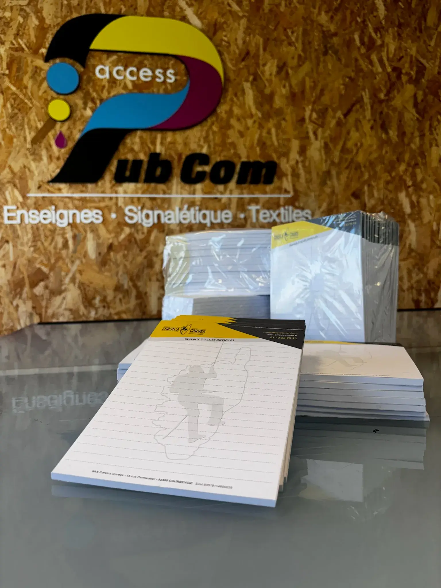 Blocs-notes à en-tête noir et jaune avec la silhouette de la Corse en filigrane, empilés devant le panneau au logo d'Access Pub Com