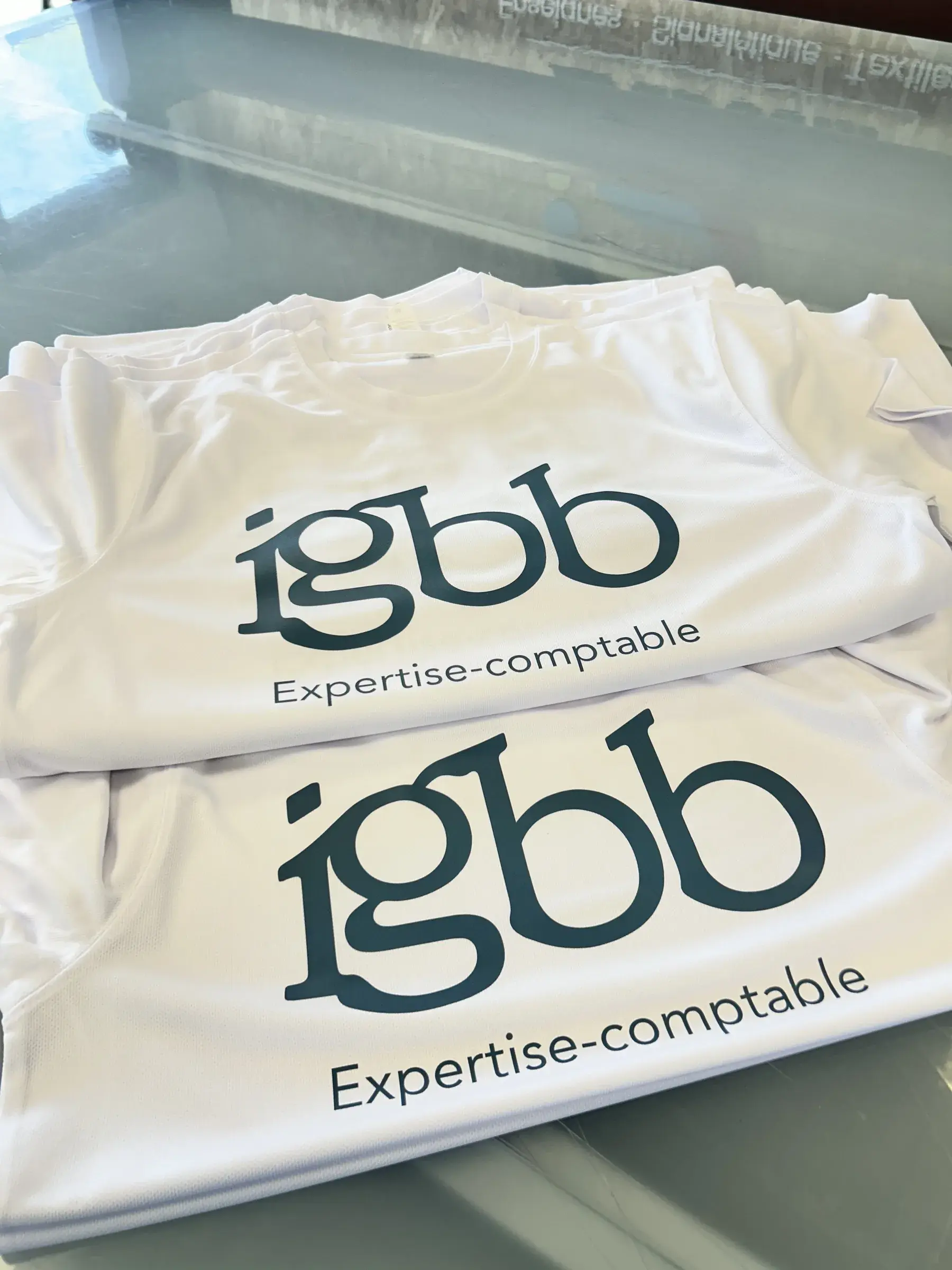Tee-shirts blancs marqués d'un logo bleu foncé, pliés en pile sur une table vitrée