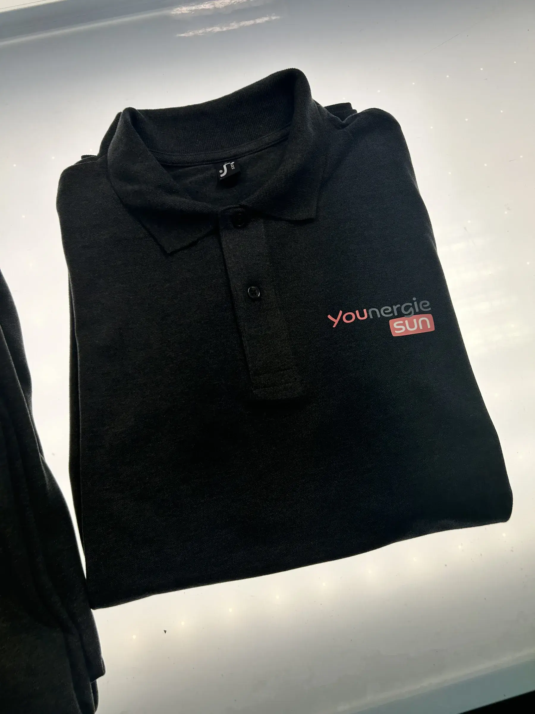 Polo anthracite brodé d'un logo rouge et gris sur la poitrine, plié sur une table lumineuse