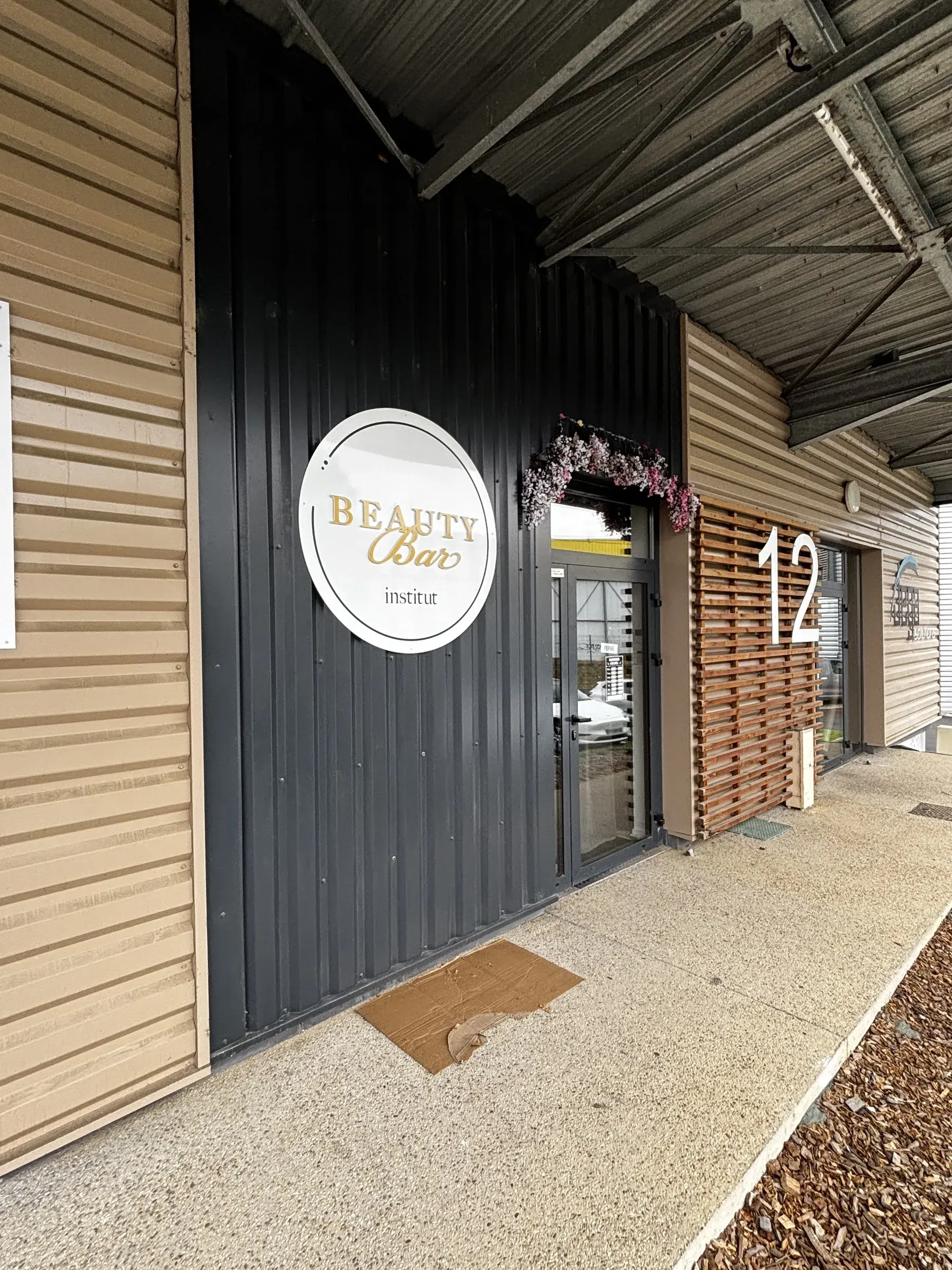 Enseigne ronde blanche à lettres dorées sur le bardage noir d'un institut, à côté d'une porte vitrée fleurie