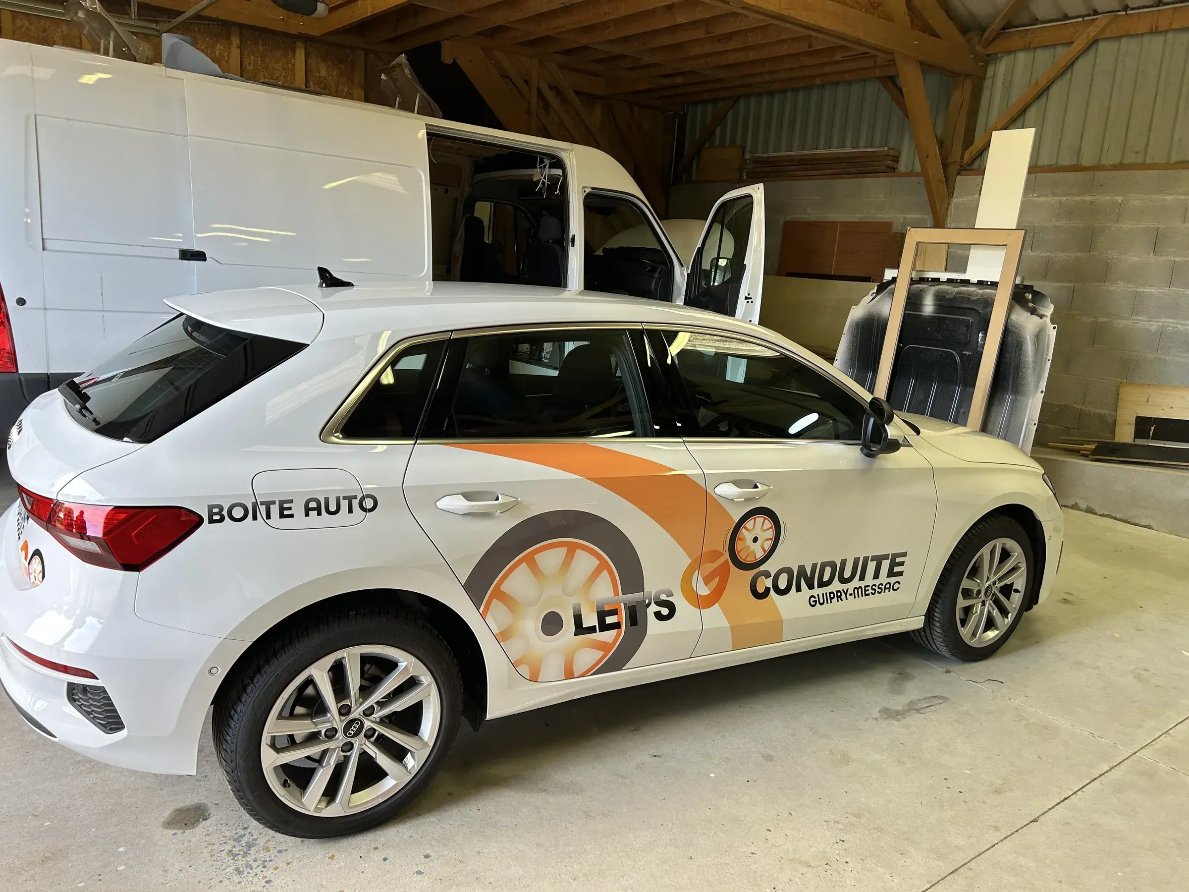 Compacte blanche marquée en orange et gris aux couleurs d'une auto-école, dans l'atelier devant un fourgon