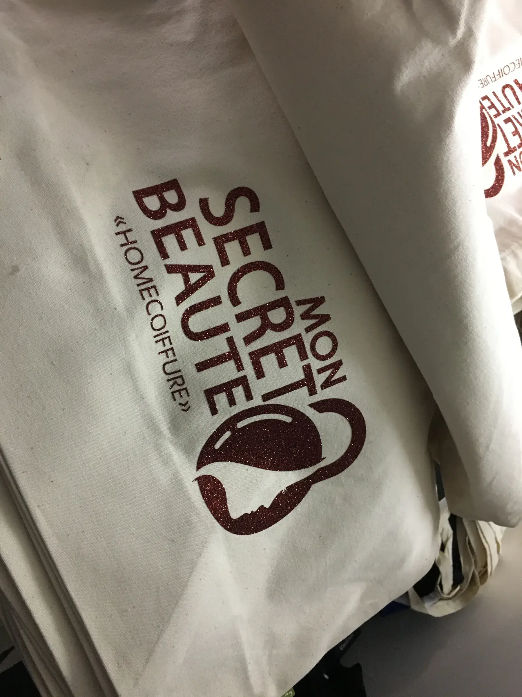 Sacs en toile écrue marqués d'un logo bordeaux pailleté, empilés les uns sur les autres