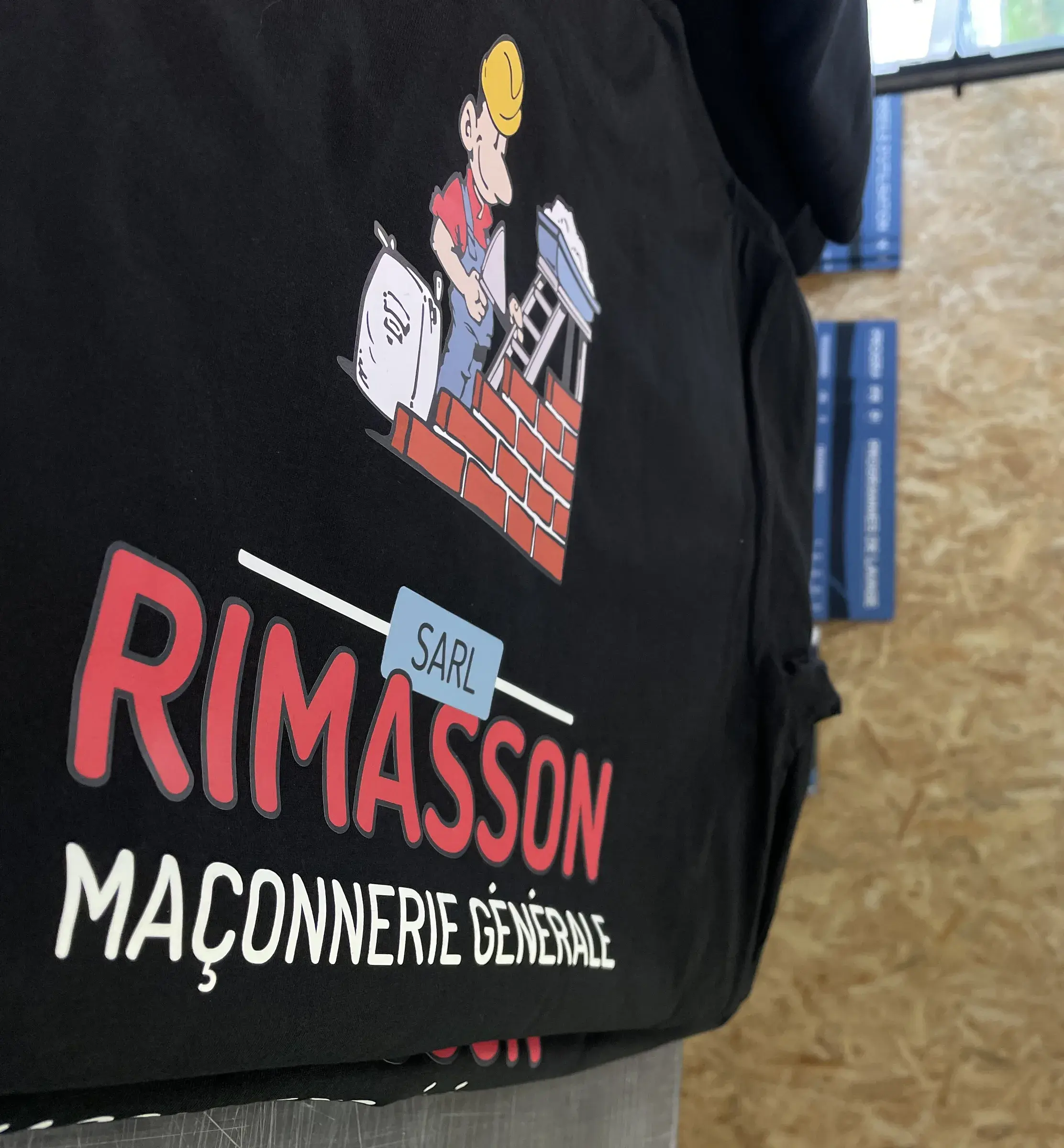 Veste noire imprimée d'une mascotte de maçon et d'un lettrage rouge et blanc, suspendue dans l'atelier