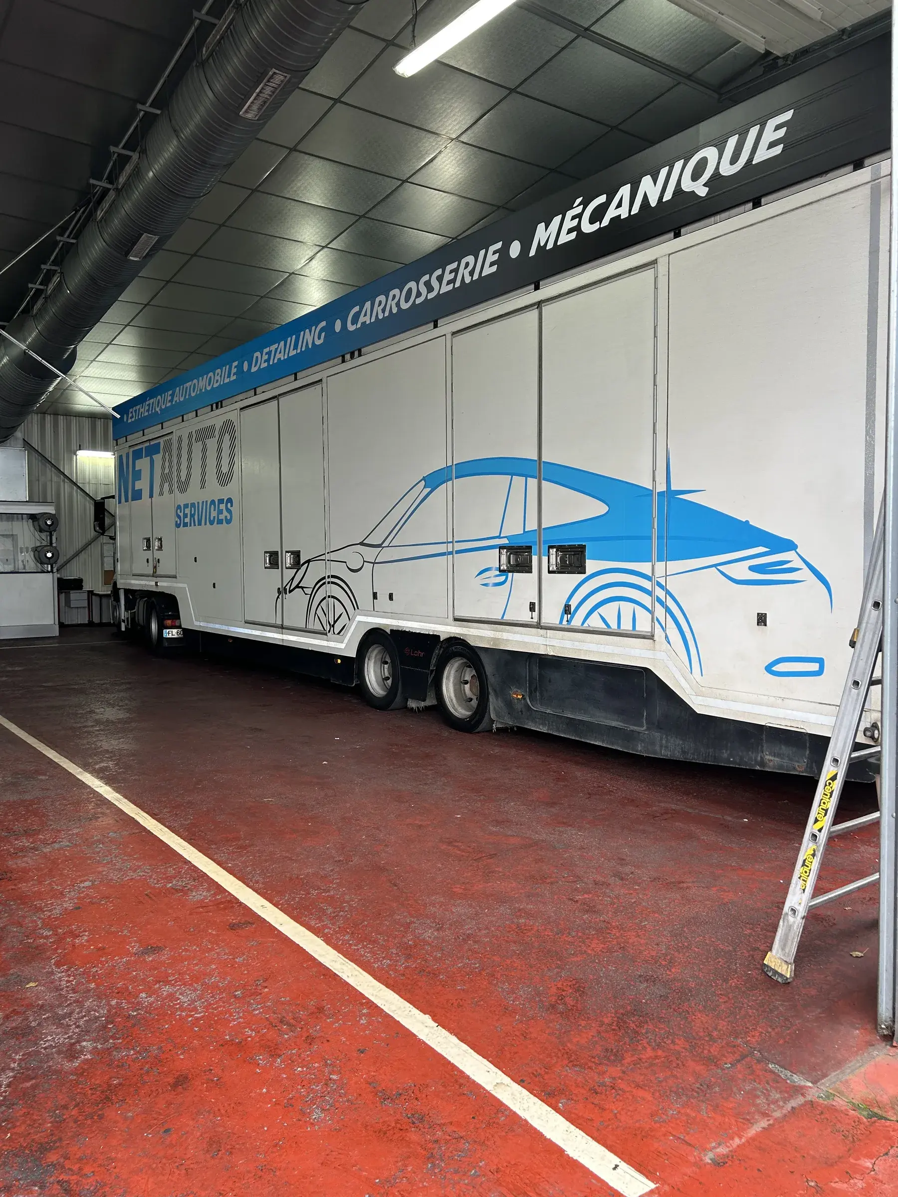 Semi-remorque blanche habillée d'une silhouette de voiture bleue et d'un bandeau de texte, dans un hangar au sol rouge