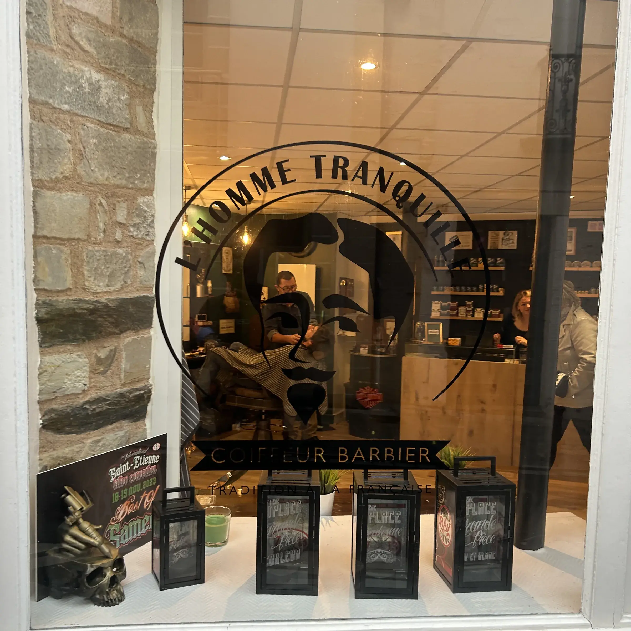Vitrine d'un salon de coiffeur barbier avec logo adhésif noir sur la vitre, lanternes et ardoise en devanture