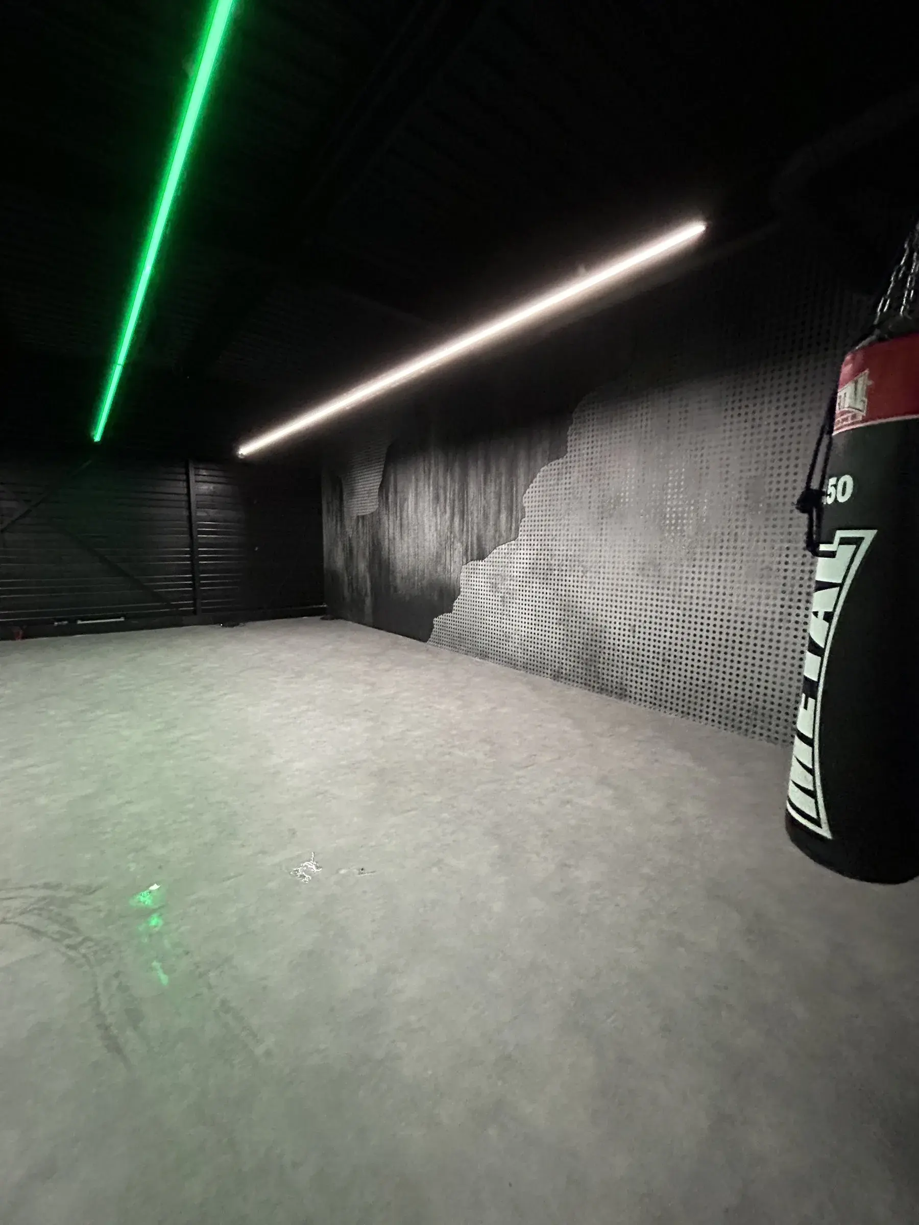 Salle d'entraînement vide avec sac de frappe, mur habillé d'un décor imprimé effet béton et métal perforé, ligne lumineuse verte au plafond