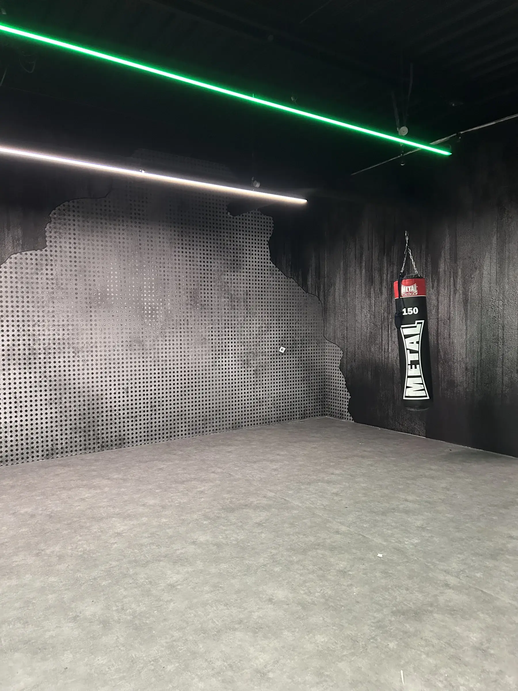 Salle de boxe aux murs noirs habillés d'un décor imprimé effet métal perforé à bord déchiré, sac de frappe suspendu et ligne lumineuse verte au plafond