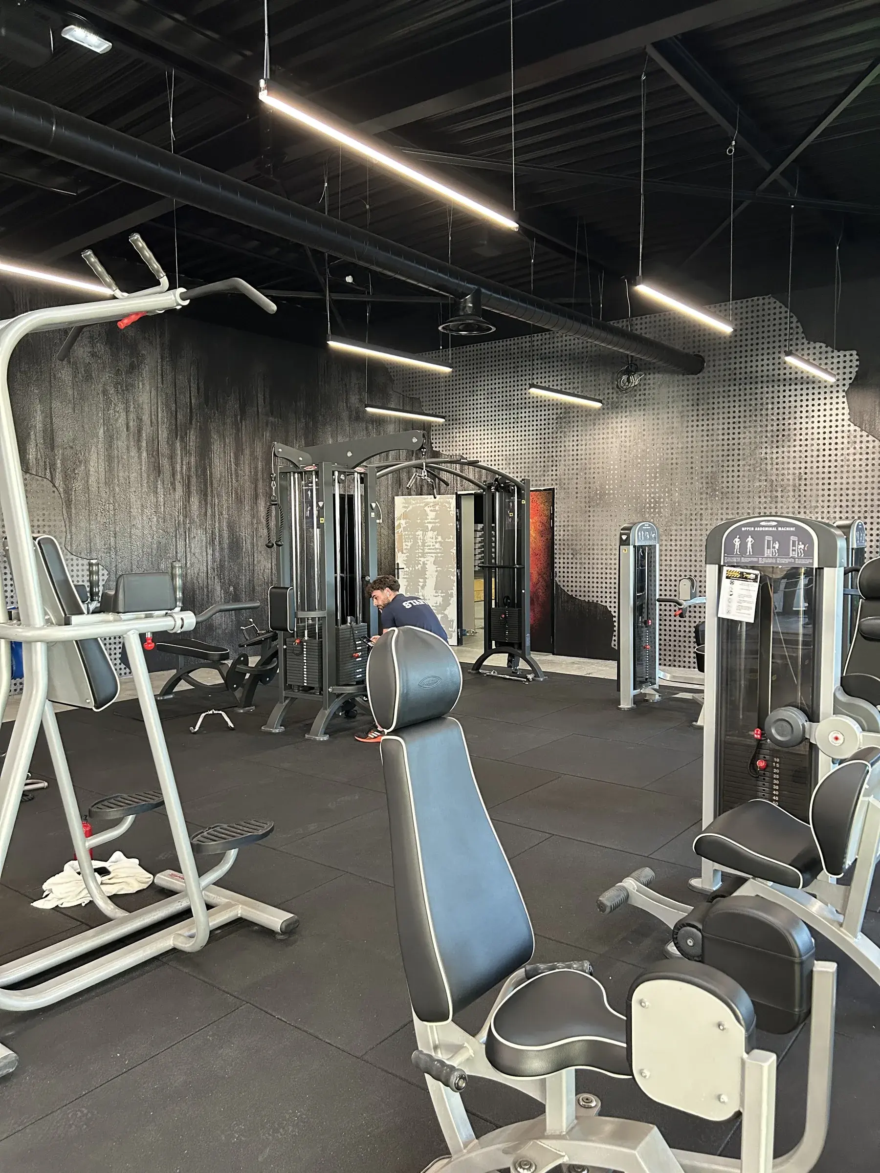 Espace musculation d'une salle de sport, murs habillés d'un décor imprimé effet béton et métal perforé sous des rampes lumineuses