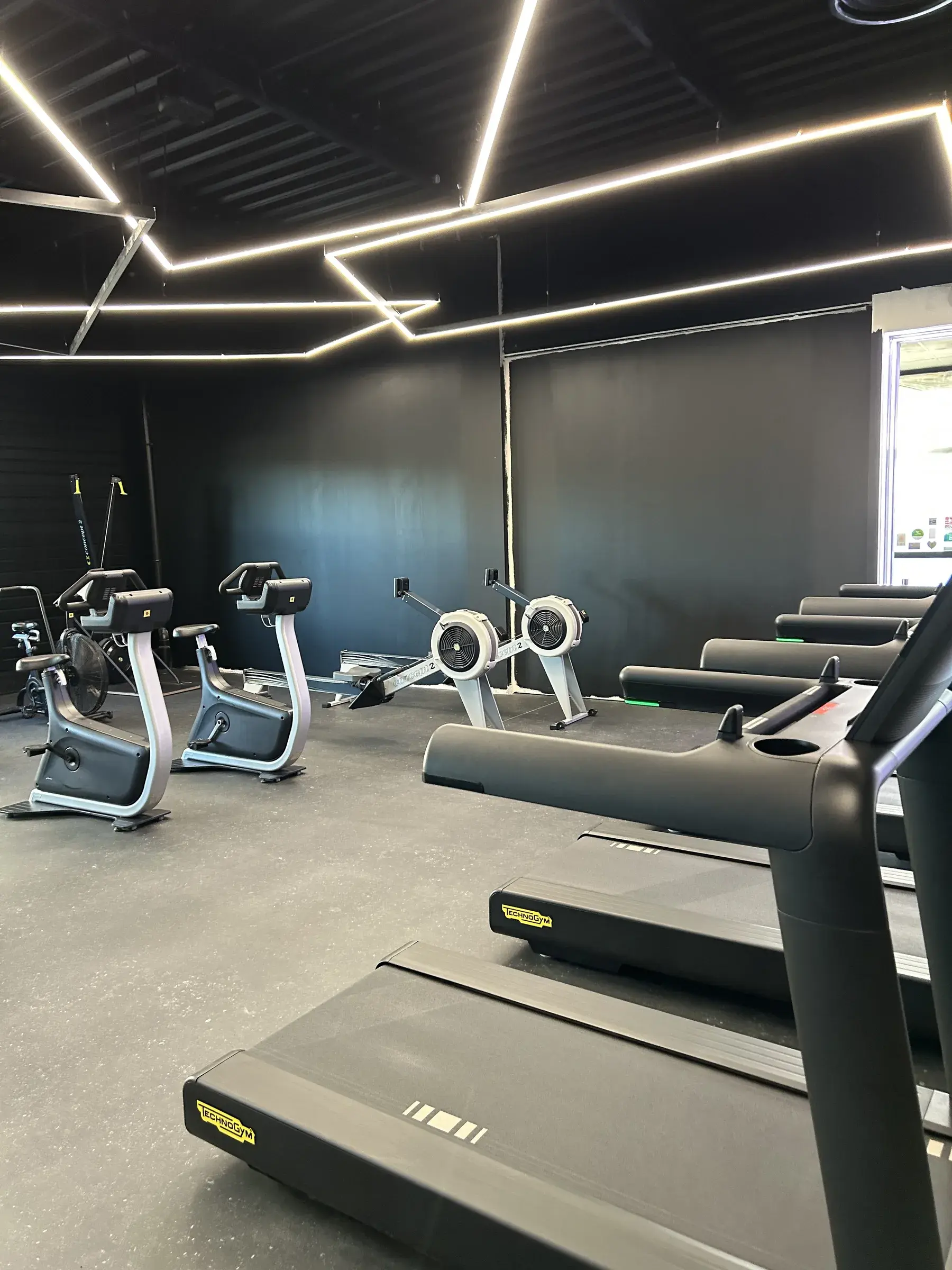 Tapis de course et vélos alignés dans une salle de sport aux murs noirs, lignes lumineuses géométriques au plafond