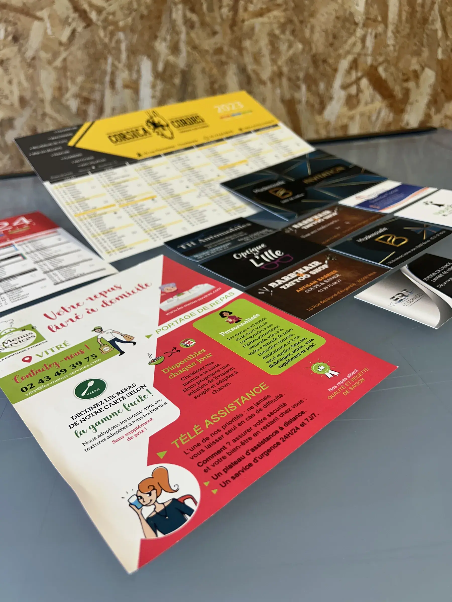 Calendrier jaune, cartes de visite noires et dépliant rouge et blanc étalés sur une table devant un mur en panneaux de bois