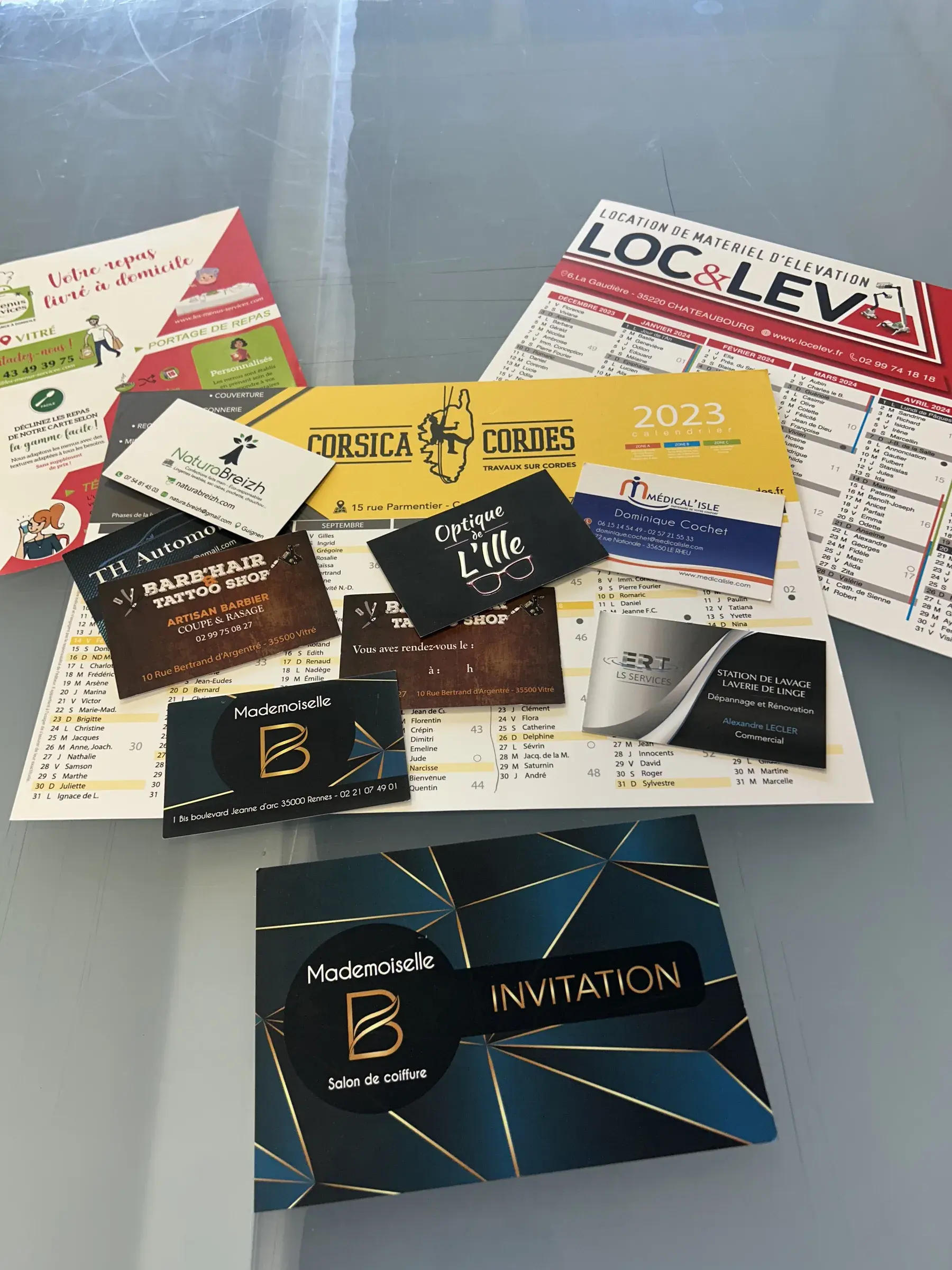 Calendriers, cartes de visite, dépliant et carton d'invitation de plusieurs clients étalés sur une table vitrée