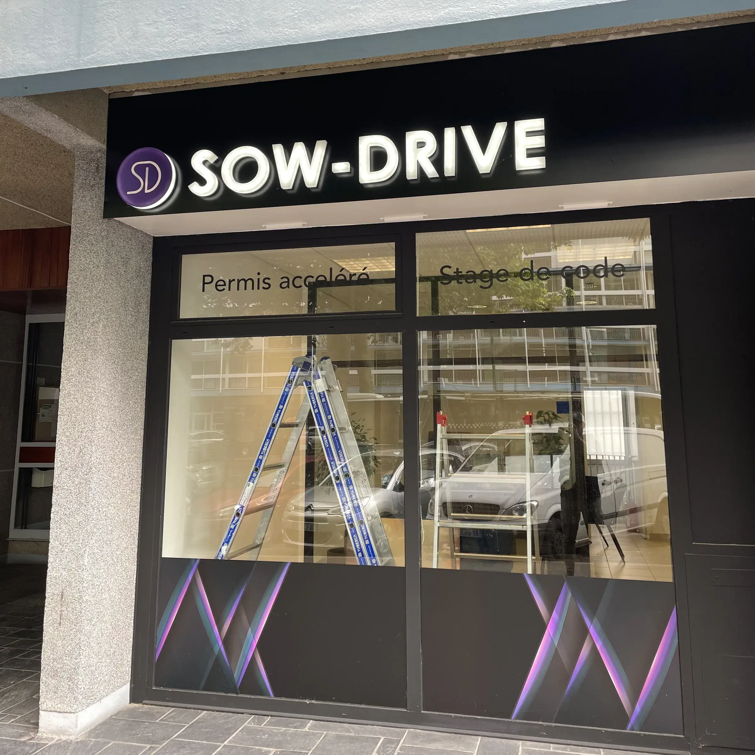 Bandeau d'enseigne noir à lettres blanches lumineuses au-dessus de la vitrine d'une auto-école, lettrage adhésif sur les vitres