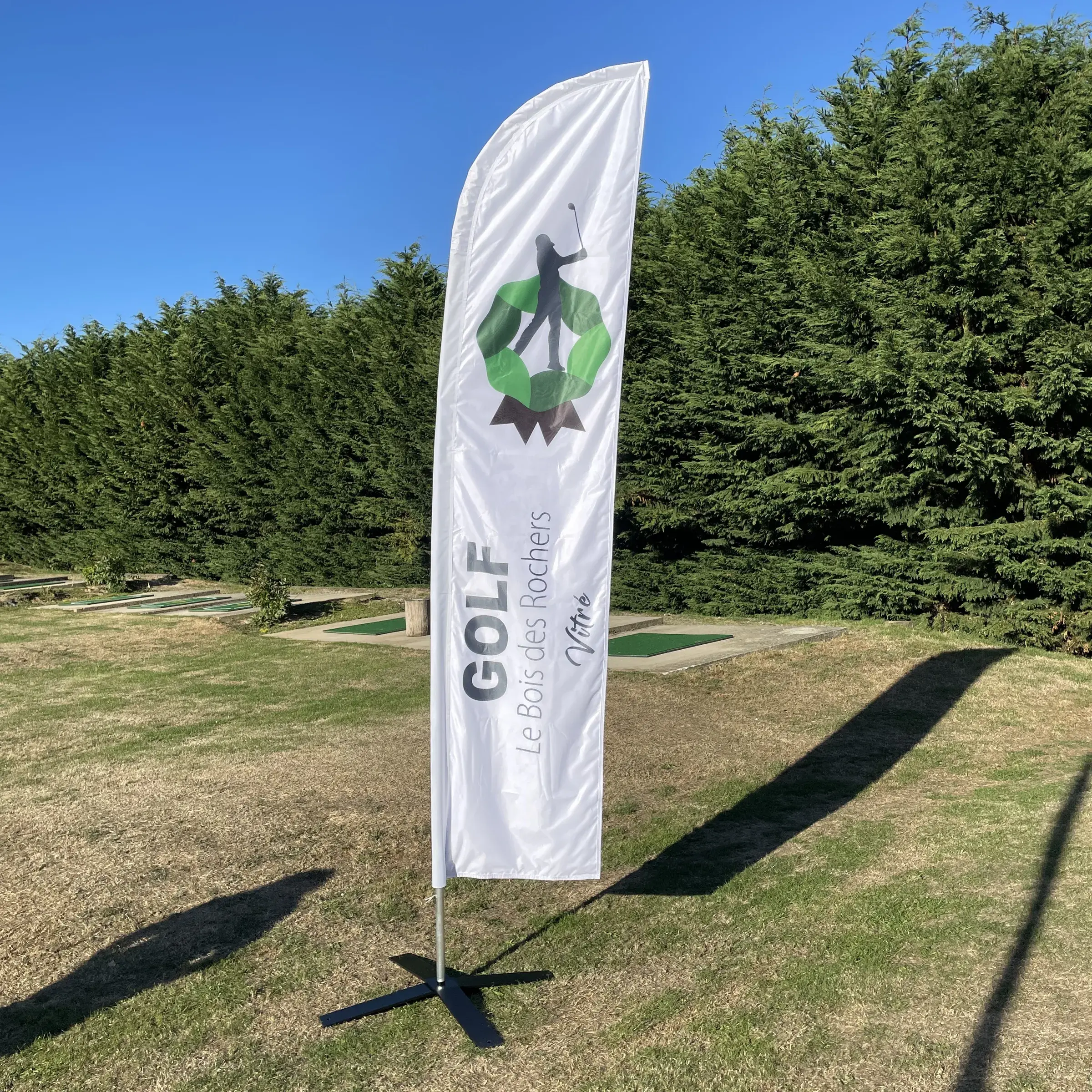 Drapeau publicitaire blanc et vert planté sur l'herbe d'un practice de golf, haie en arrière-plan