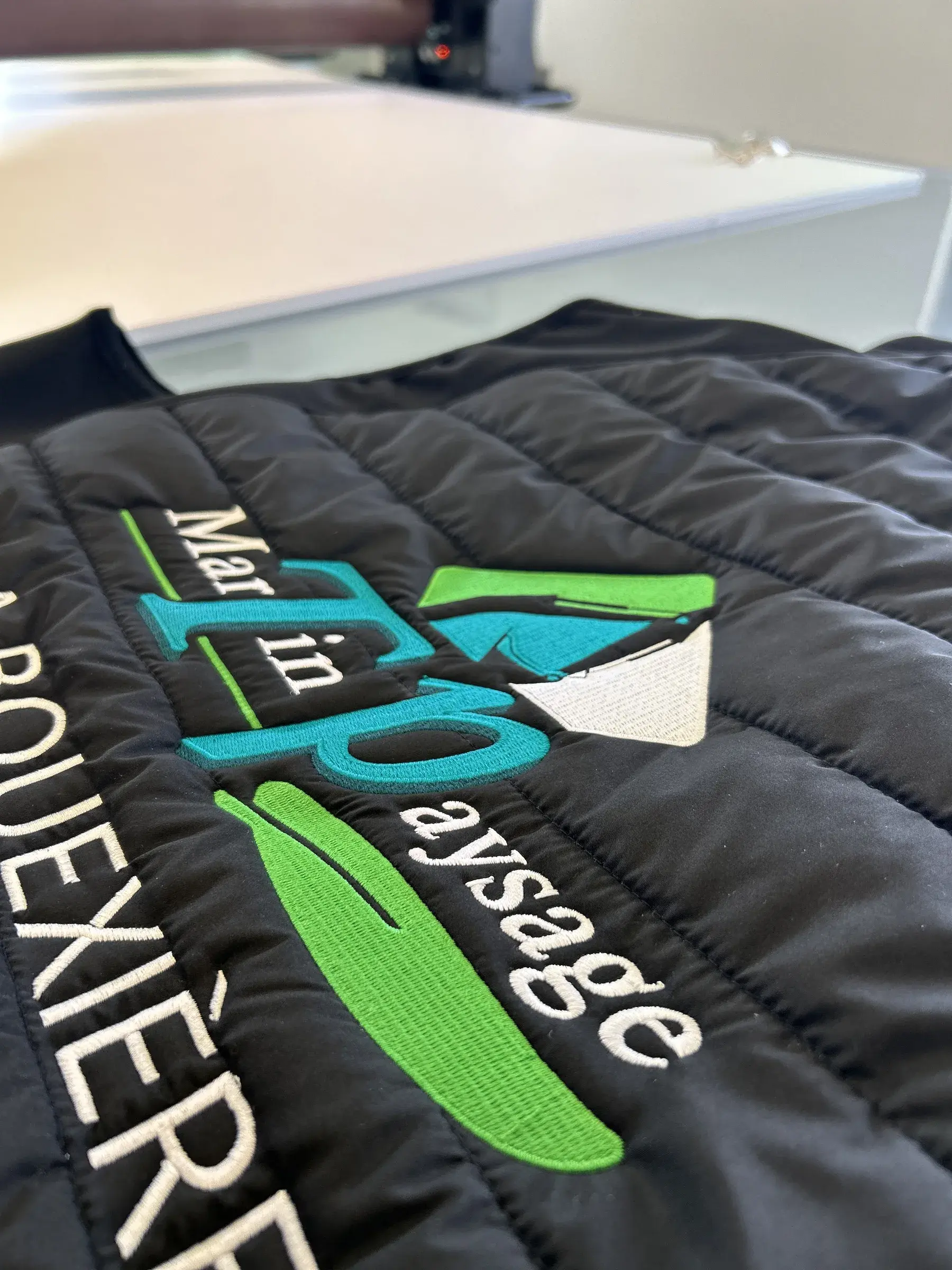 Broderie verte, bleue et blanche en cours sur une veste matelassée noire, devant la machine à broder