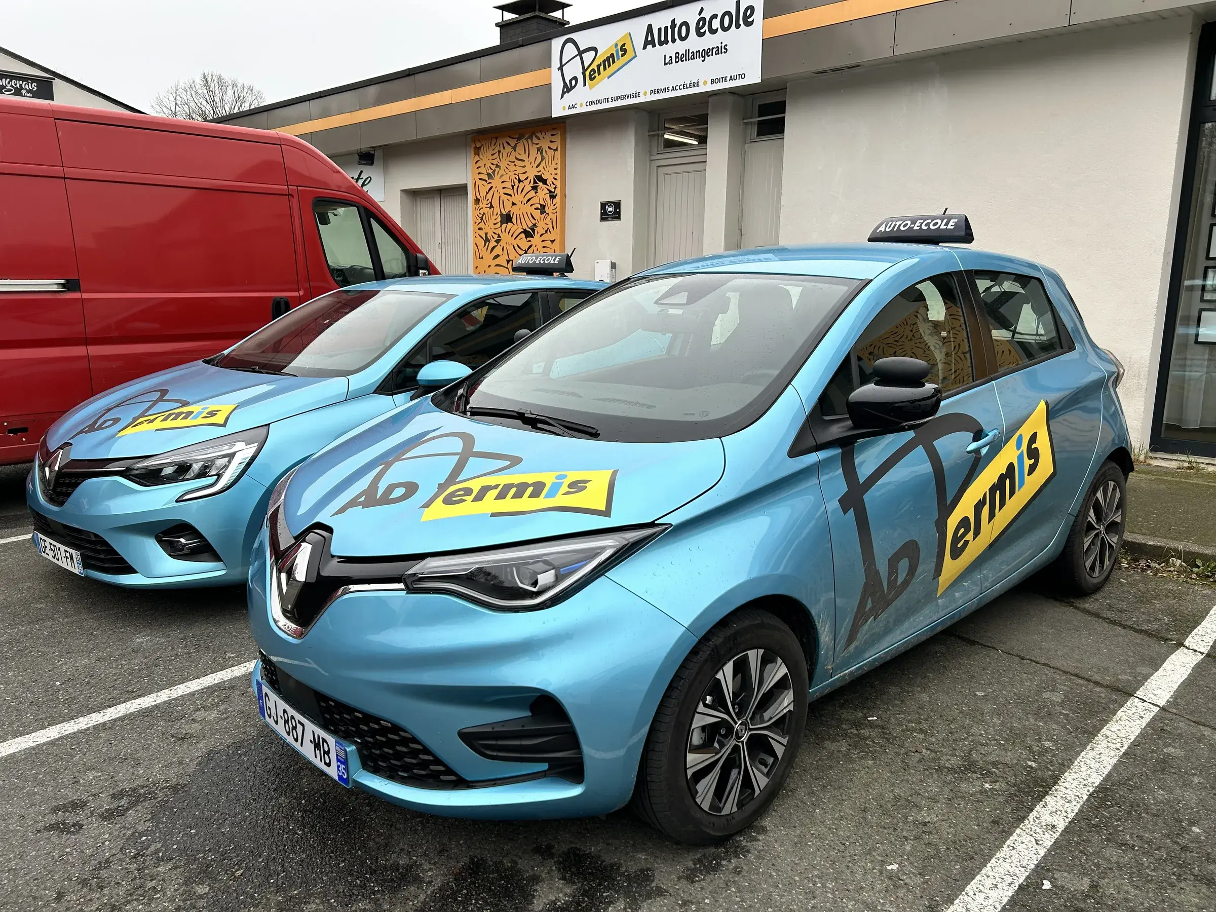 Deux citadines bleu clair marquées au logo d'une auto-école, garées devant la façade de l'agence