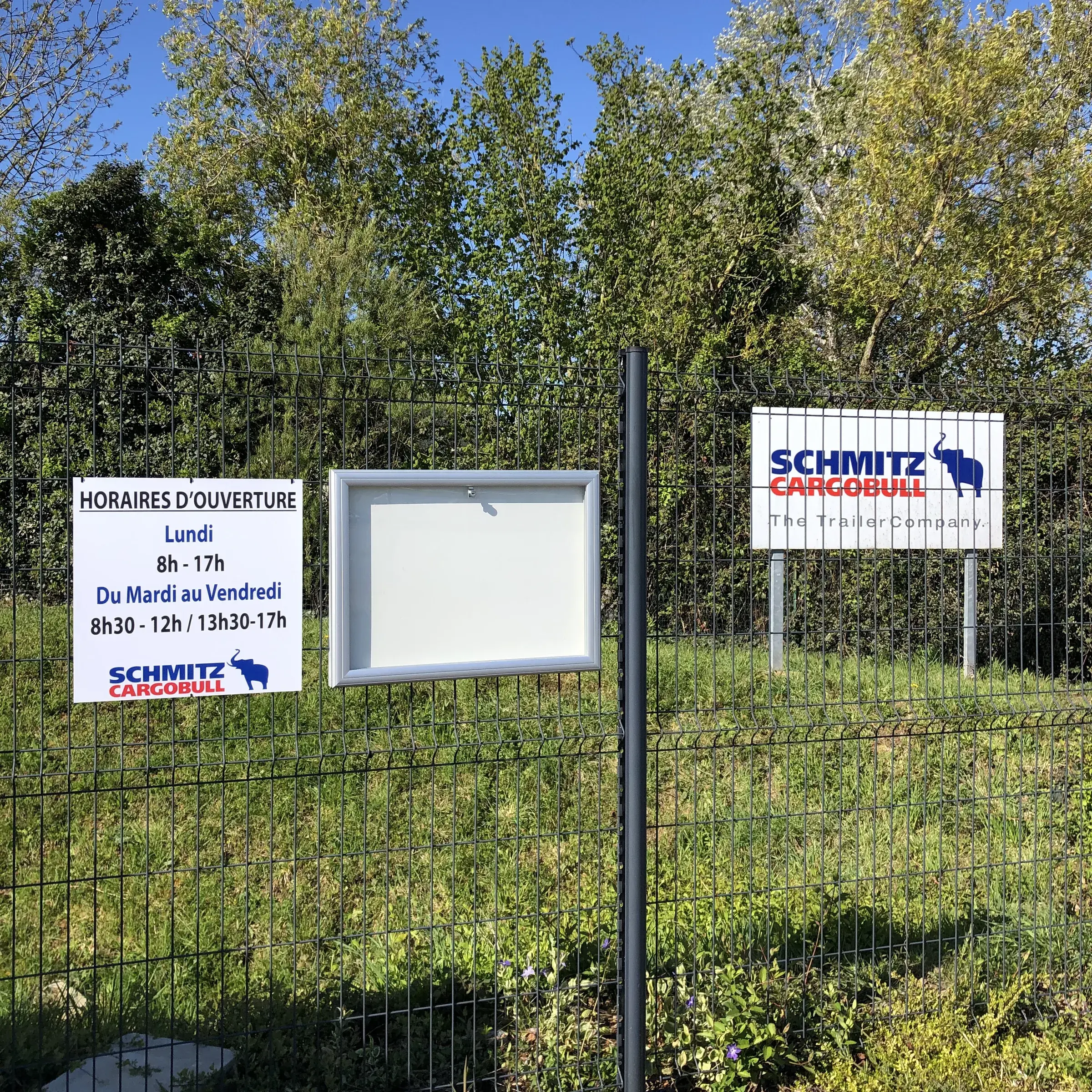 Panneau d'horaires et vitrine d'affichage fixés sur une clôture grillagée, panneau d'entreprise sur poteaux en arrière-plan