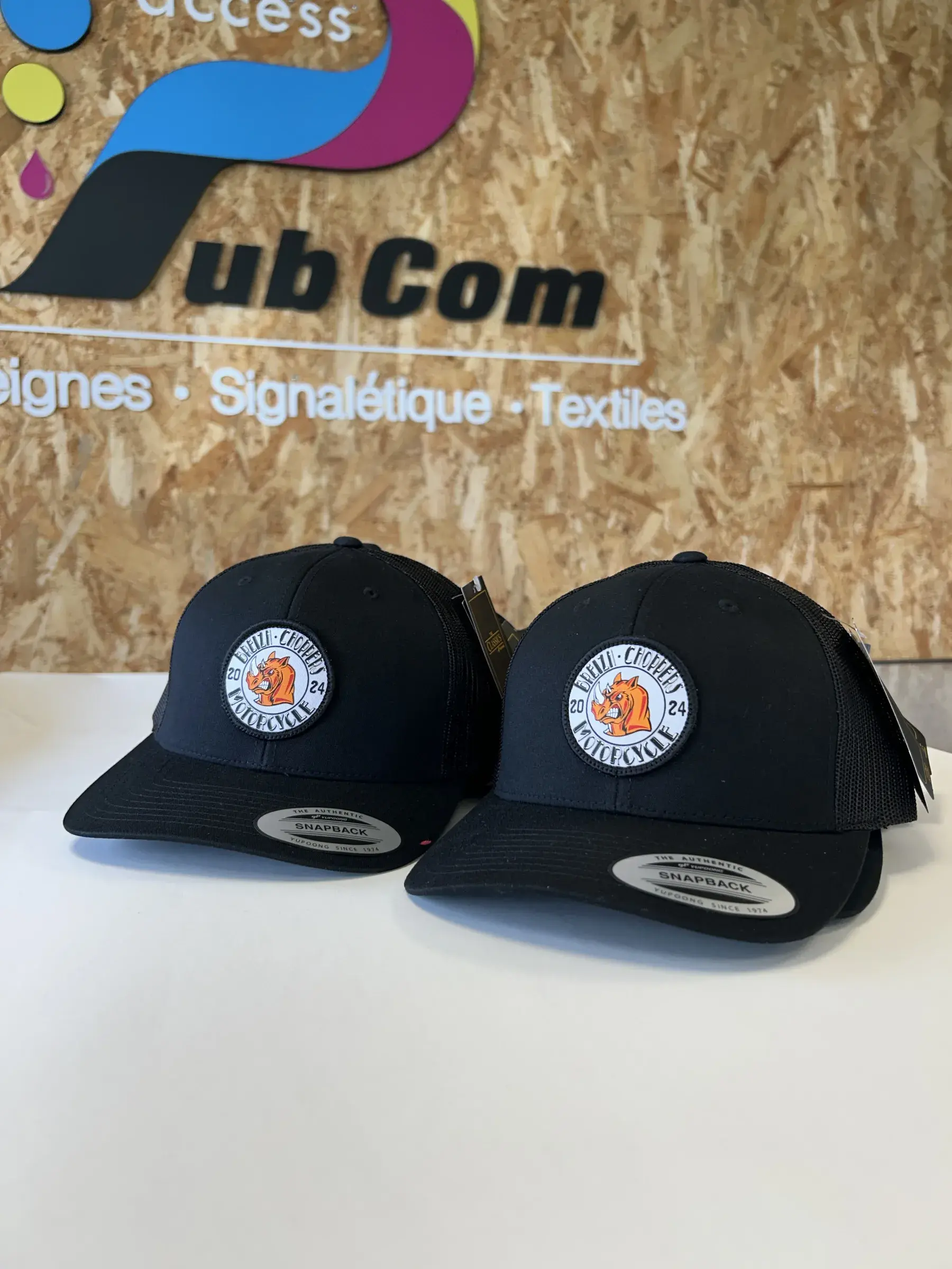 Deux casquettes noires à écusson rond brodé, posées devant le panneau au logo d'Access Pub Com
