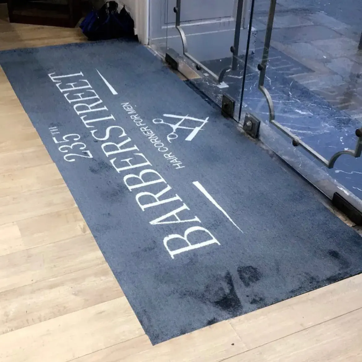 Tapis d'entrée gris imprimé au logo d'un salon de coiffure, devant une porte vitrée