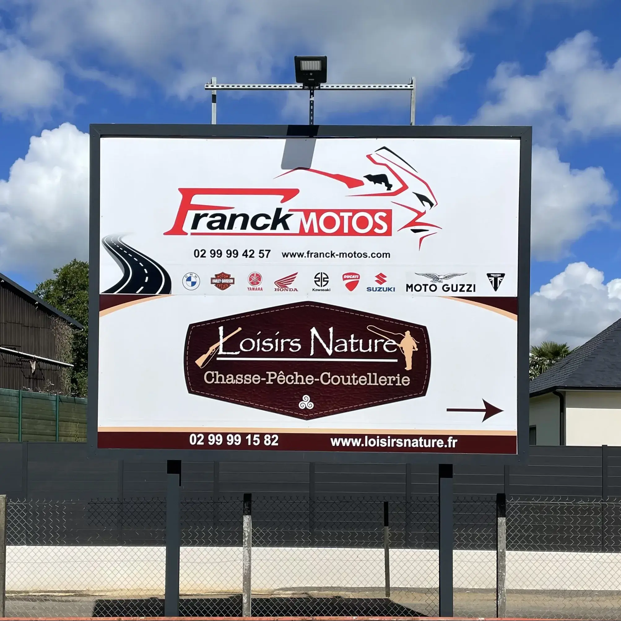 Grand panneau publicitaire sur poteaux partagé entre deux commerces, projecteur en tête, devant une clôture